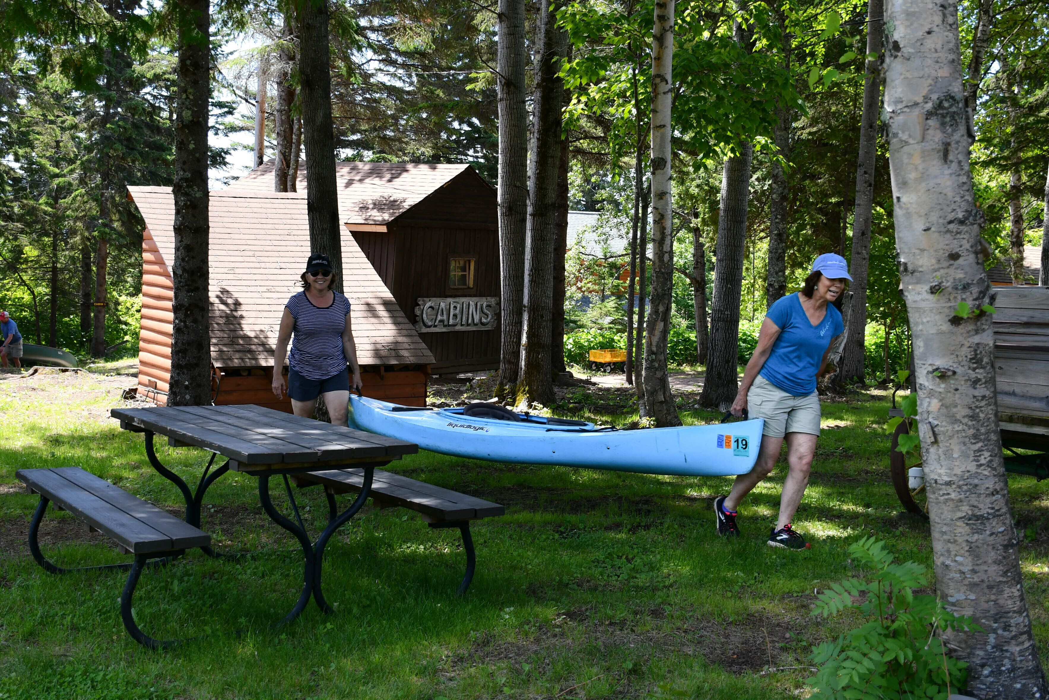 Una cabaña de madera con la leyenda 'CABINS' en un área boscosa, con mesas de picnic y una canoa.