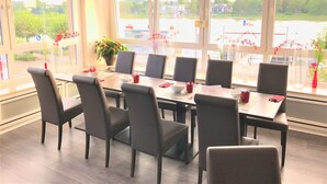 Ein Konferenzraum mit großem Tisch und Stühlen, großen Fenstern mit Blick auf einen Fluss und Gebäude, sowie Blumentöpfen auf dem Fensterbrett.