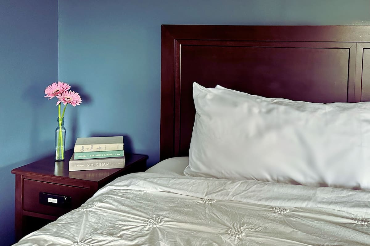 A neatly made bed with white linens, a wooden nightstand with a vase of pink flowers, and a stack of books.
