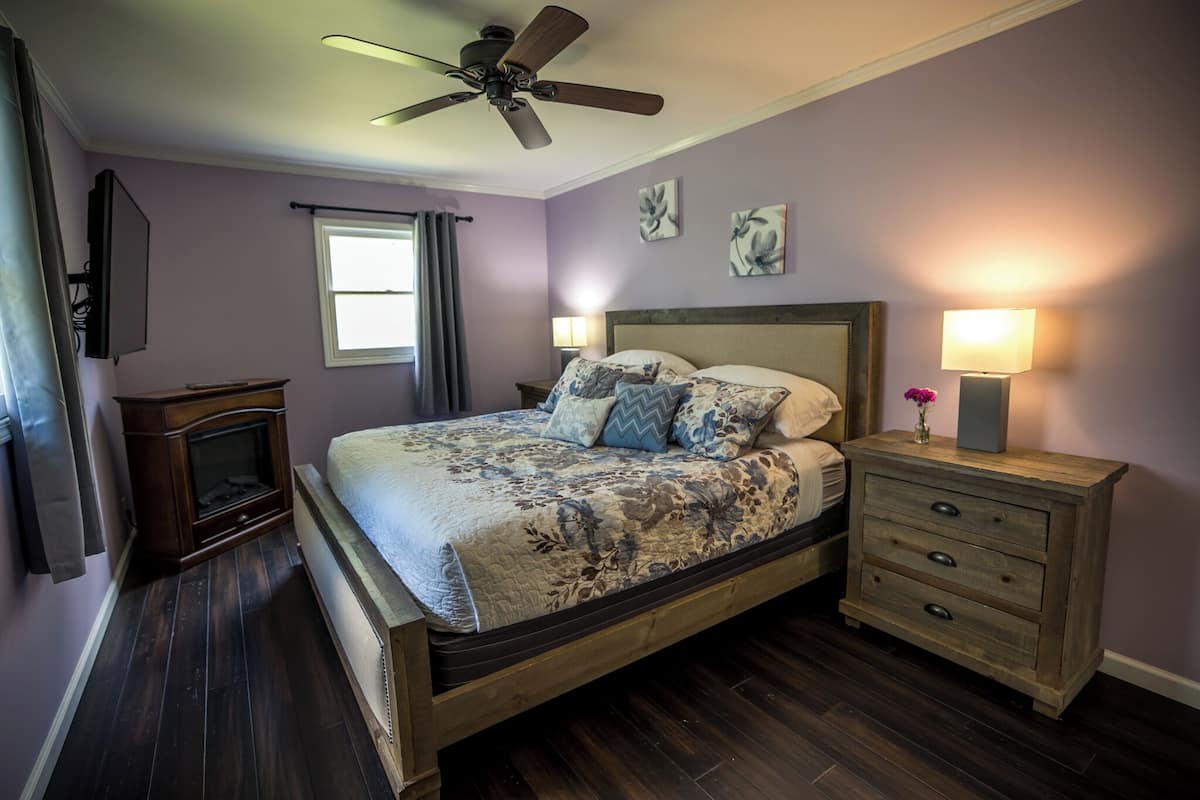 A bedroom with a bed, a nightstand, a ceiling fan, a window with curtains, and a fireplace.