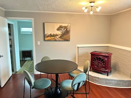 Un comedor con una mesa redonda y cuatro sillas, una chimenea con estufa roja, y un cuadro en la pared.