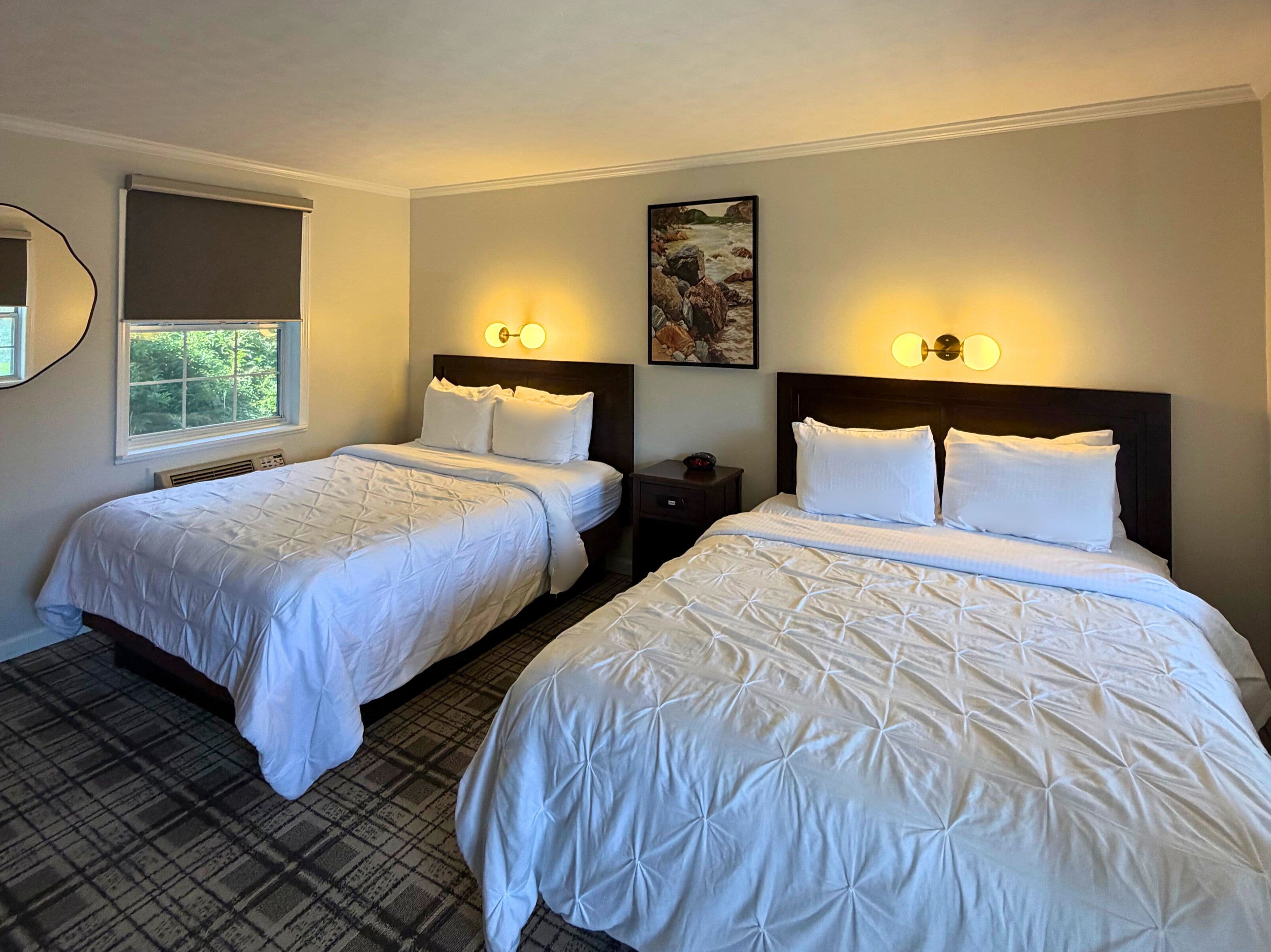 A hotel room with two beds, a window, a painting, and wall sconces.