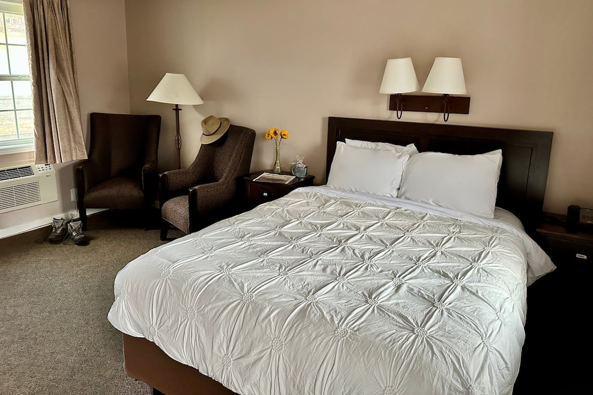 A hotel room with a large bed, two bedside lamps, a chair, a small table with a vase of flowers, and a window with curtains.