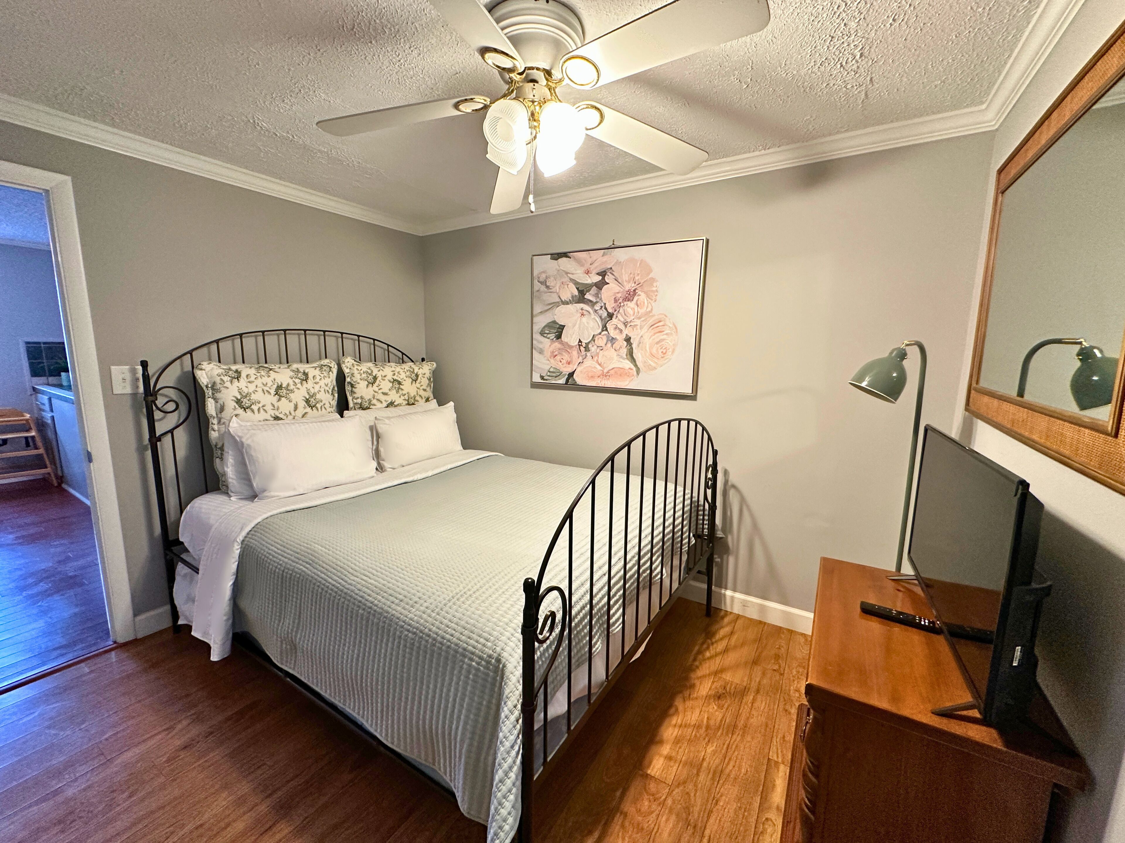 A bedroom with a bed, a desk, a chair, a desk lamp, a ceiling fan, and a framed picture on the wall.