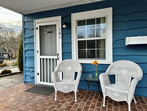 A blue house with a white door, two wicker chairs, a small table with a vase of flowers, and a window.