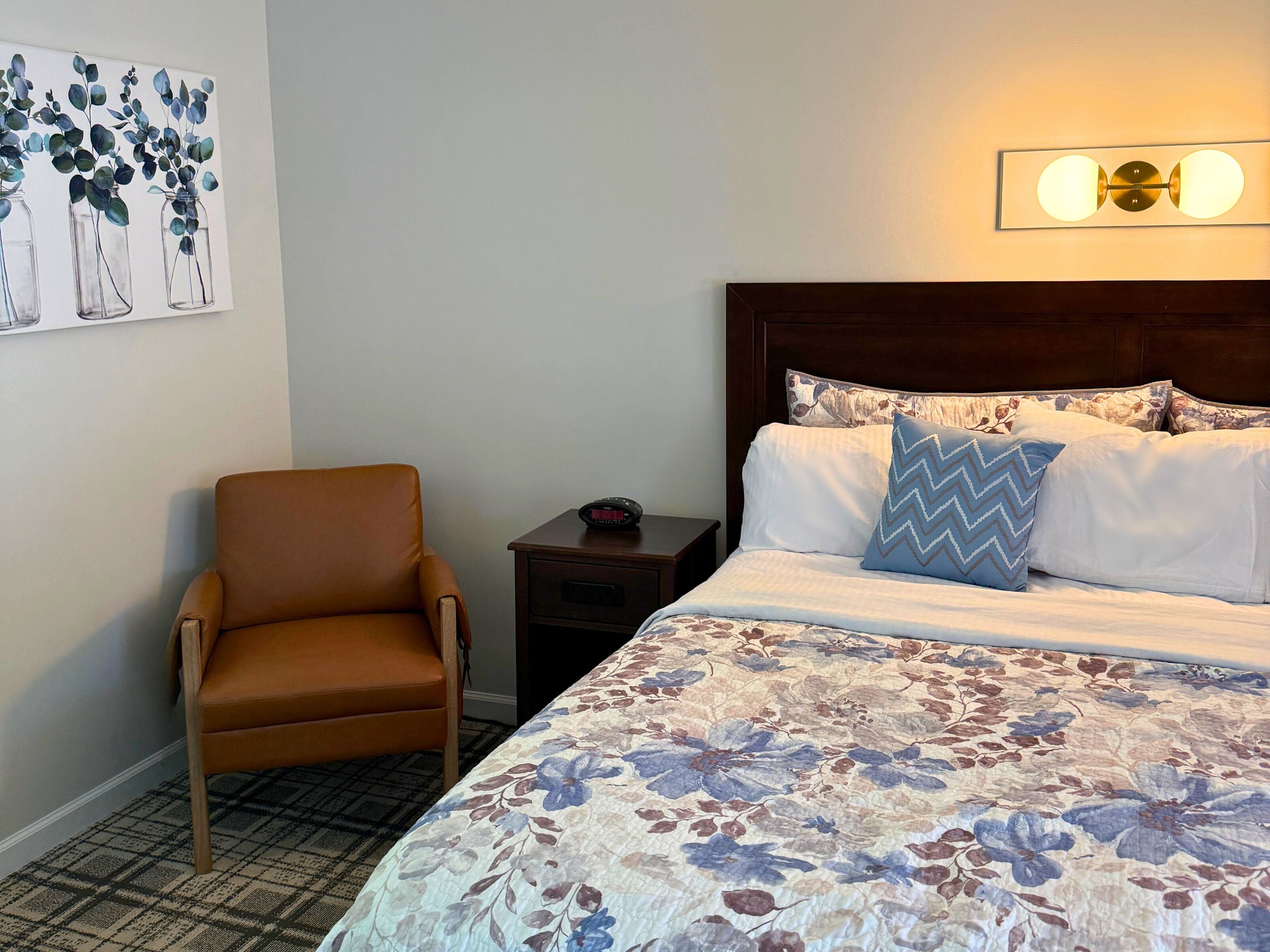 A hotel room with a bed, a chair, a nightstand, and a wall-mounted light fixture.