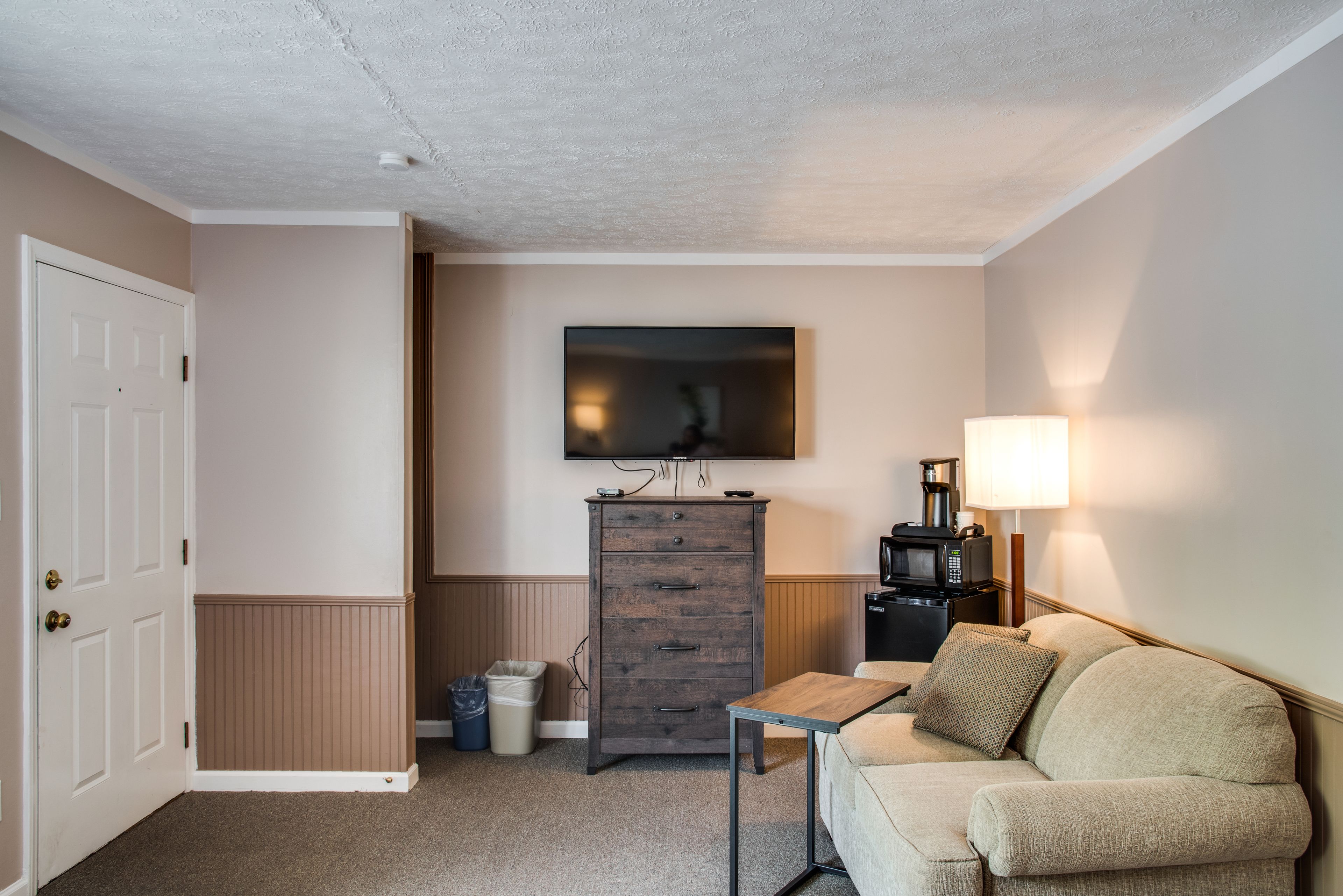 A small, well-lit room with a beige sofa, a wooden cabinet, a small table, a TV mounted on the wall, and a lamp on a side table.