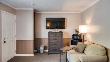 A small, well-lit room with a beige sofa, a wooden cabinet, a small table, a TV mounted on the wall, and a lamp on a side table.