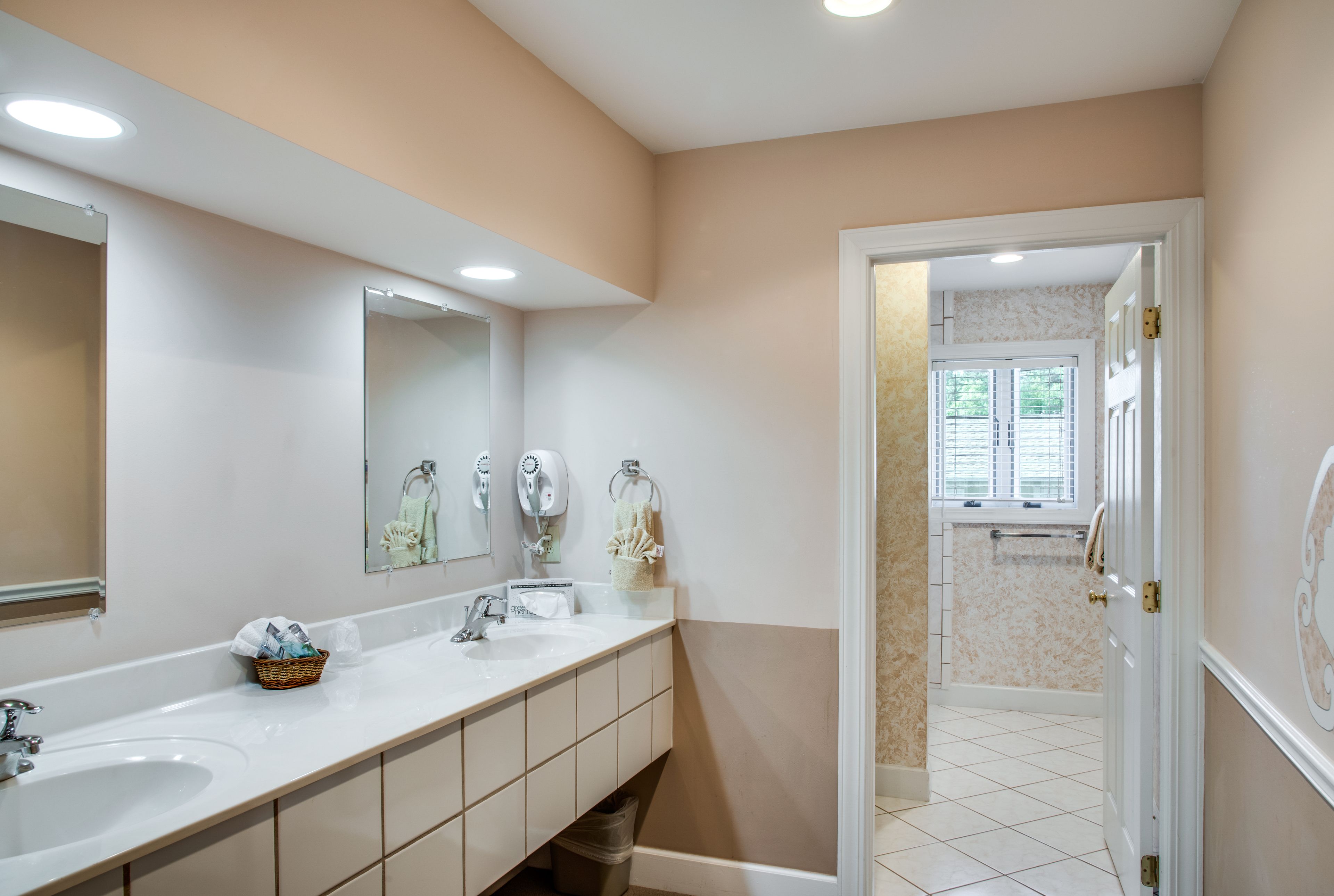 Une salle de bain moderne avec un grand lavabo, un miroir et deux sèche-cheveux. On y trouve une poubelle sous le lavabo et une fenêtre donnant sur l’espace de la douche.