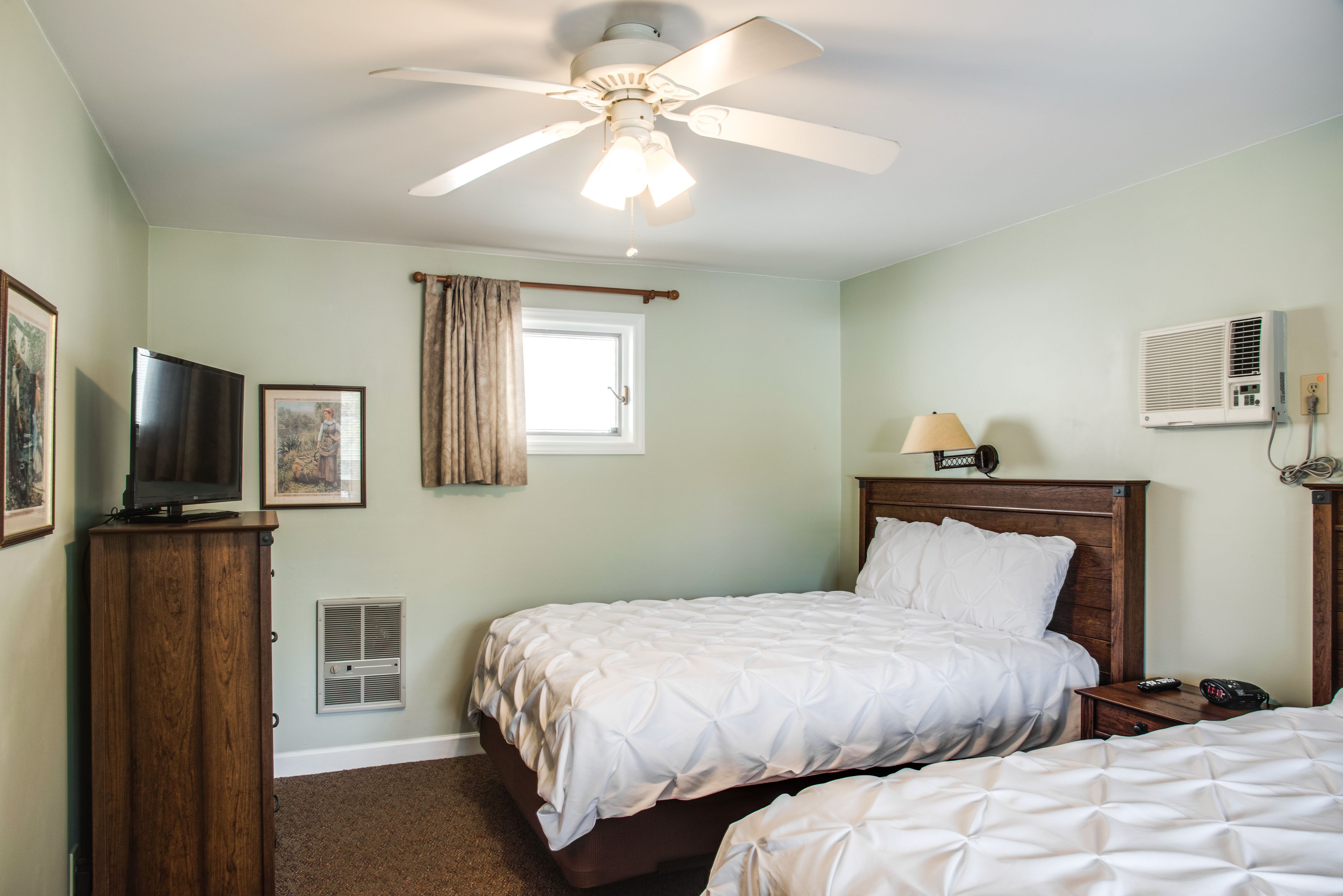Une chambre d’hôtel avec deux lits, un ventilateur de plafond, un téléviseur posé sur une commode en bois, une fenêtre avec des rideaux et un appareil de climatisation.