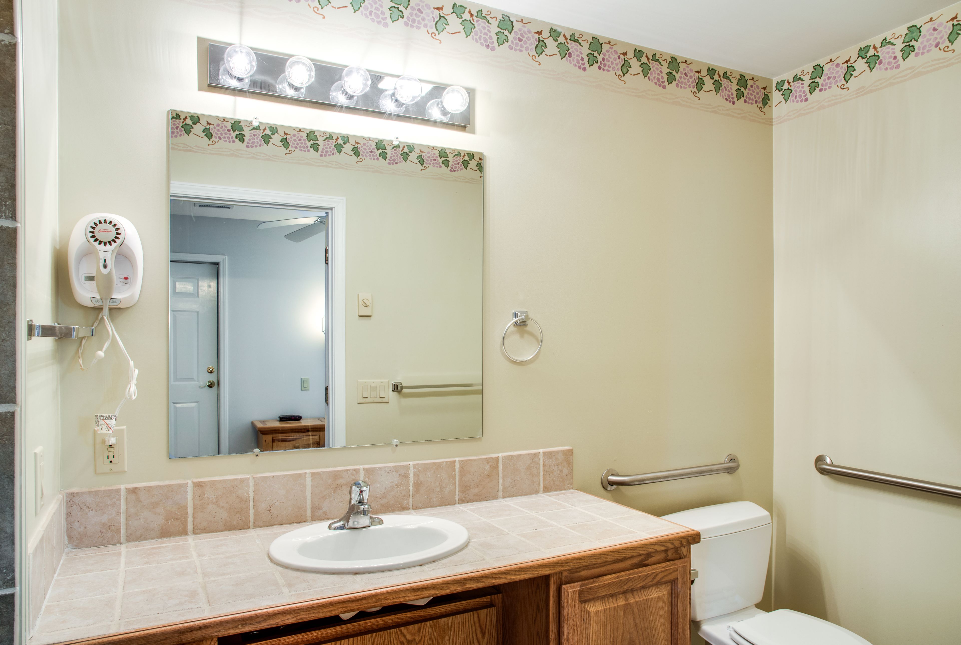 Baño con lavamanos, espejo e inodoro. Hay un secamanos en la pared y un perchero para toallas.