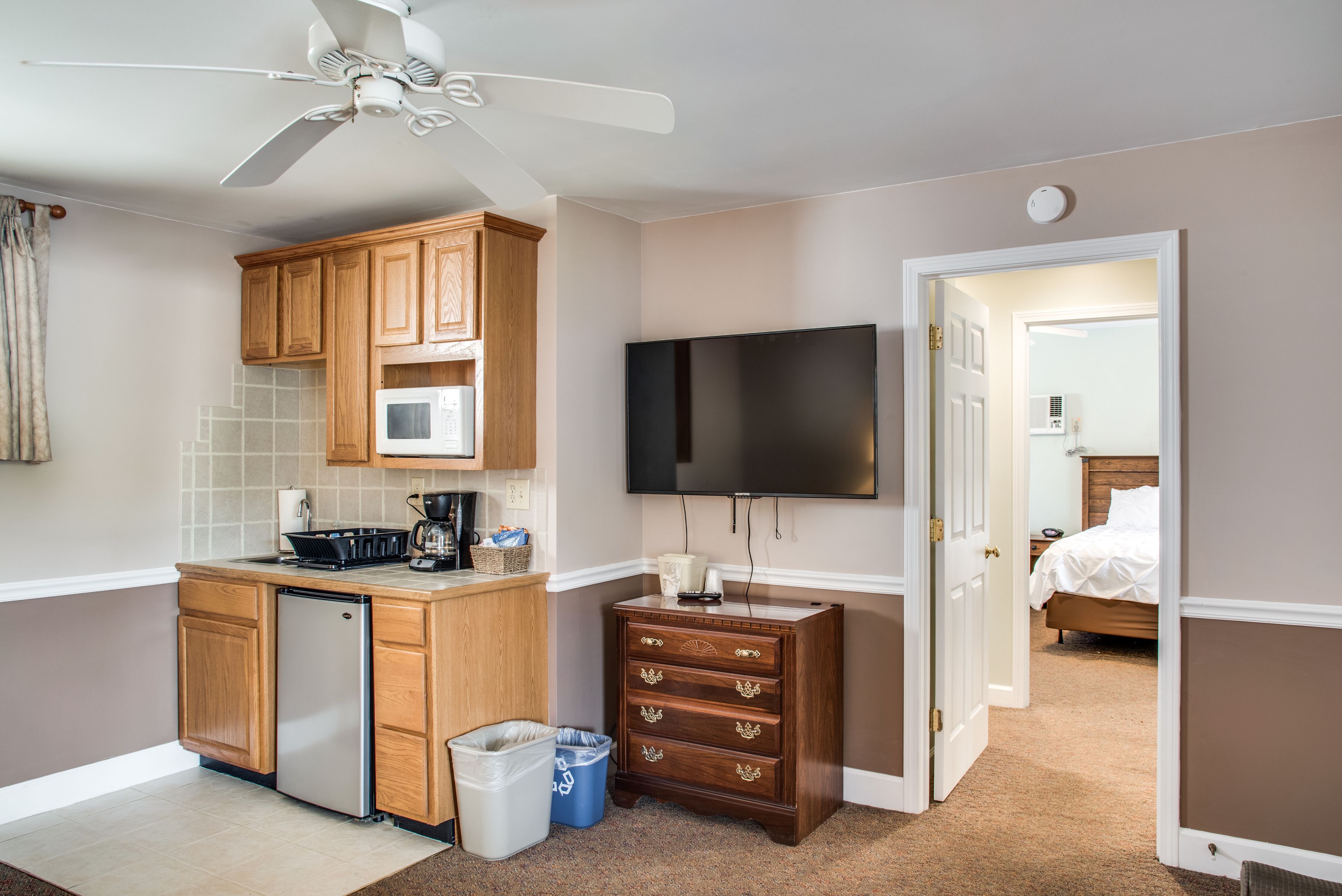 Une chambre d’hôtel compacte comprenant un coin cuisine, un ventilateur de plafond, un téléviseur à écran plat et un lit.