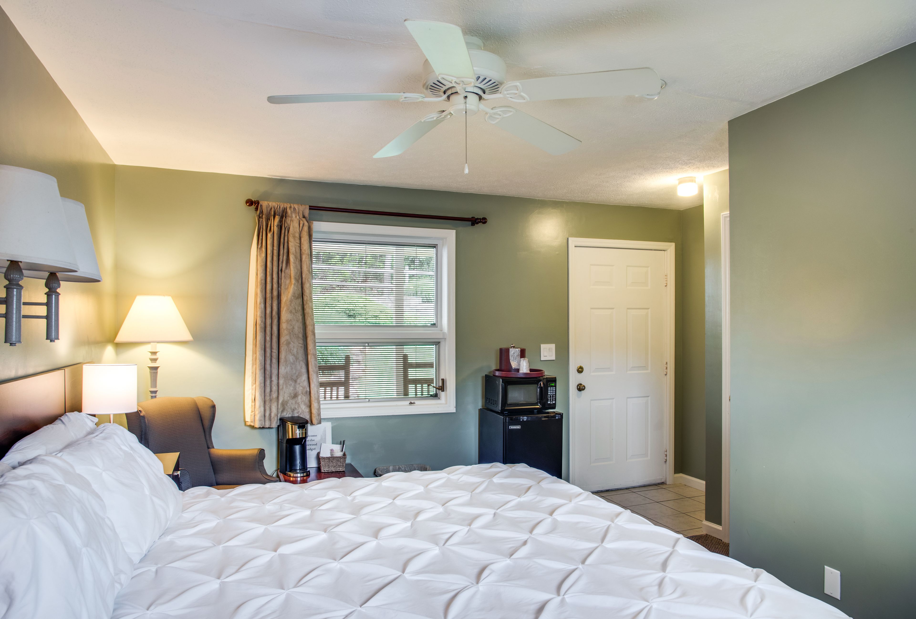 A hotel room with a bed, a ceiling fan, a window with curtains, a door, and a small table with a lamp.