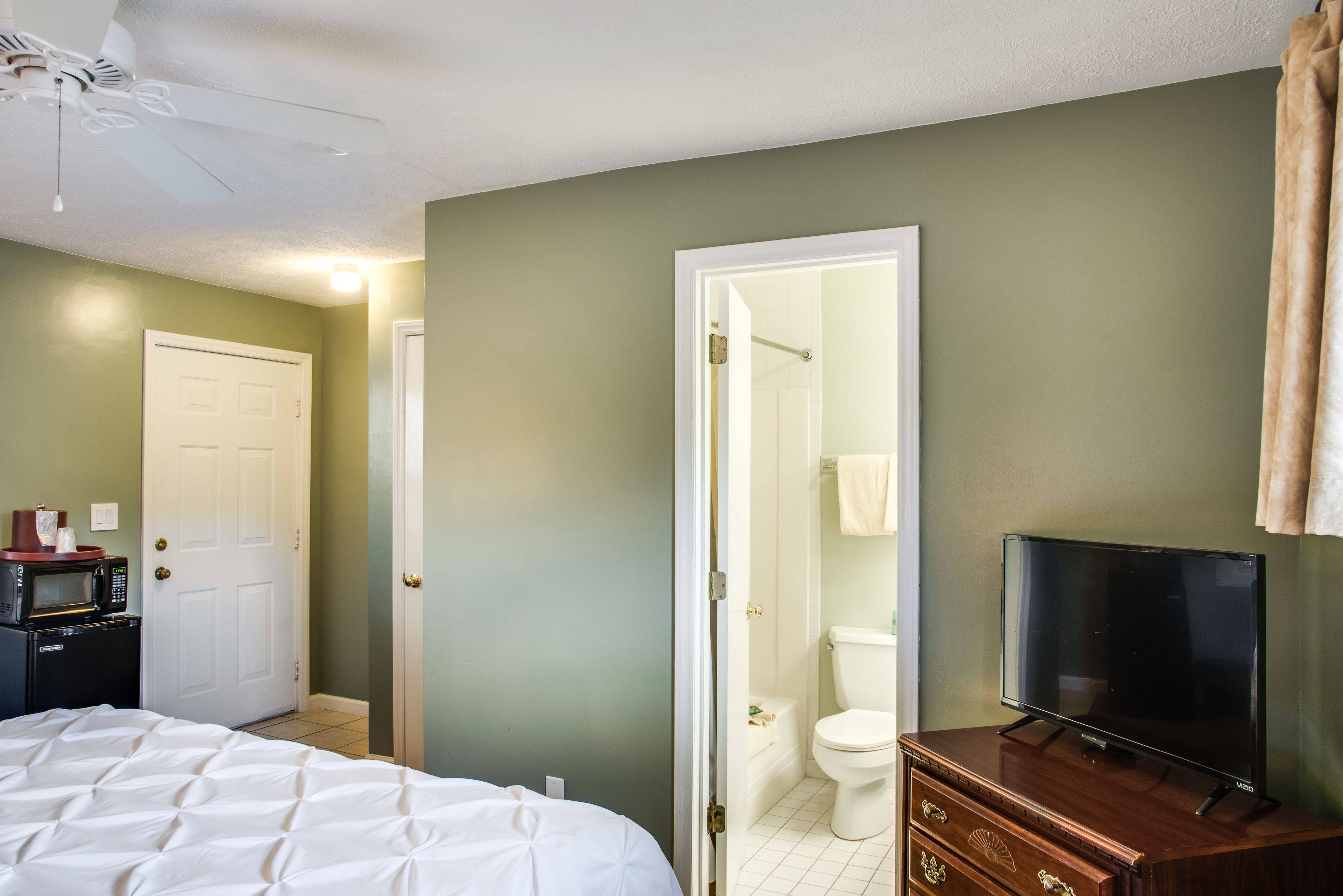 A hotel room with a bed, a TV on a wooden dresser, a microwave, and a door leading to a bathroom.