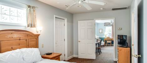 A bedroom with a bed, a ceiling fan, a TV stand, and a door leading to a dining area.