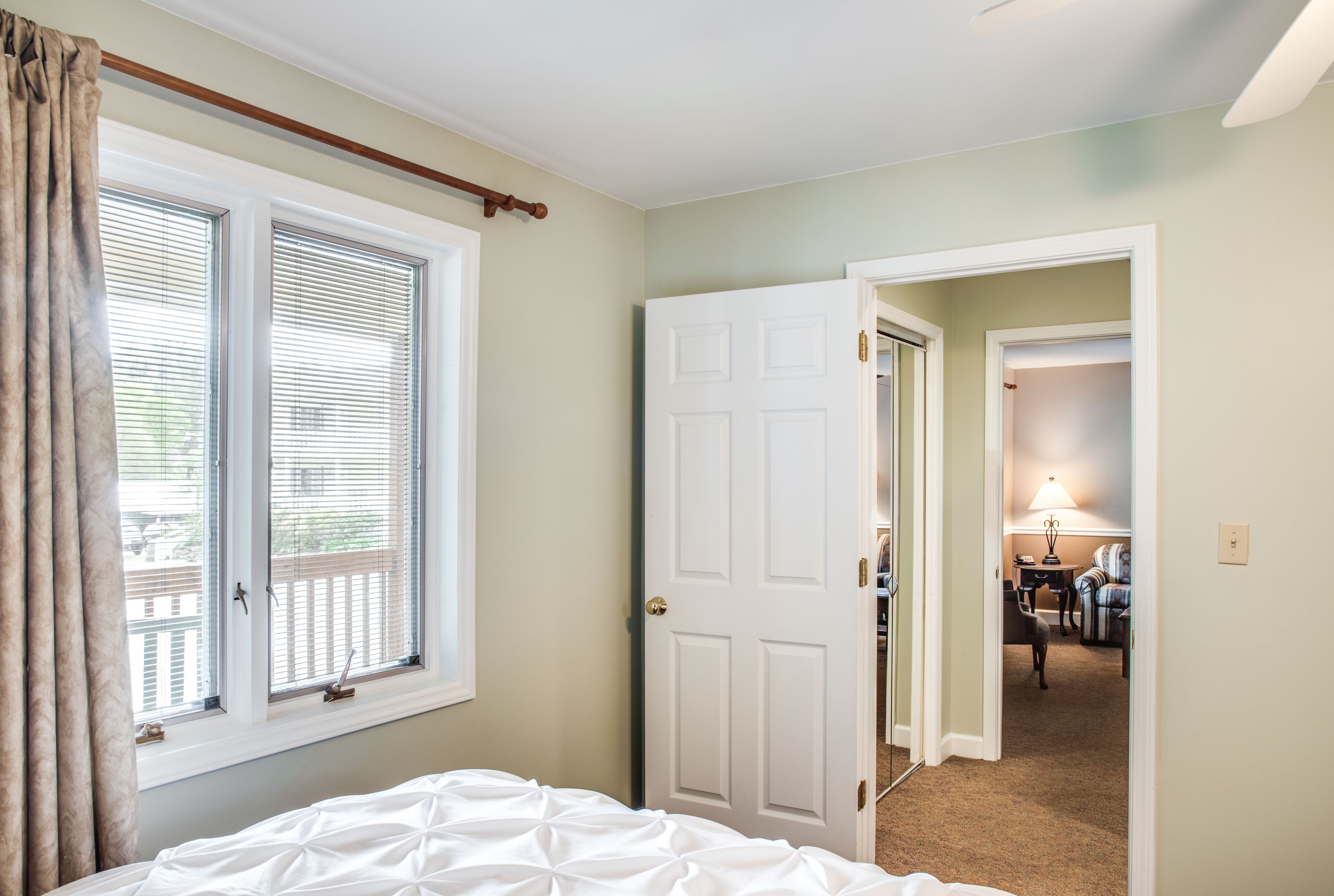 A bedroom with a bed, a window with blinds, a door leading to another room, and a visible hallway.