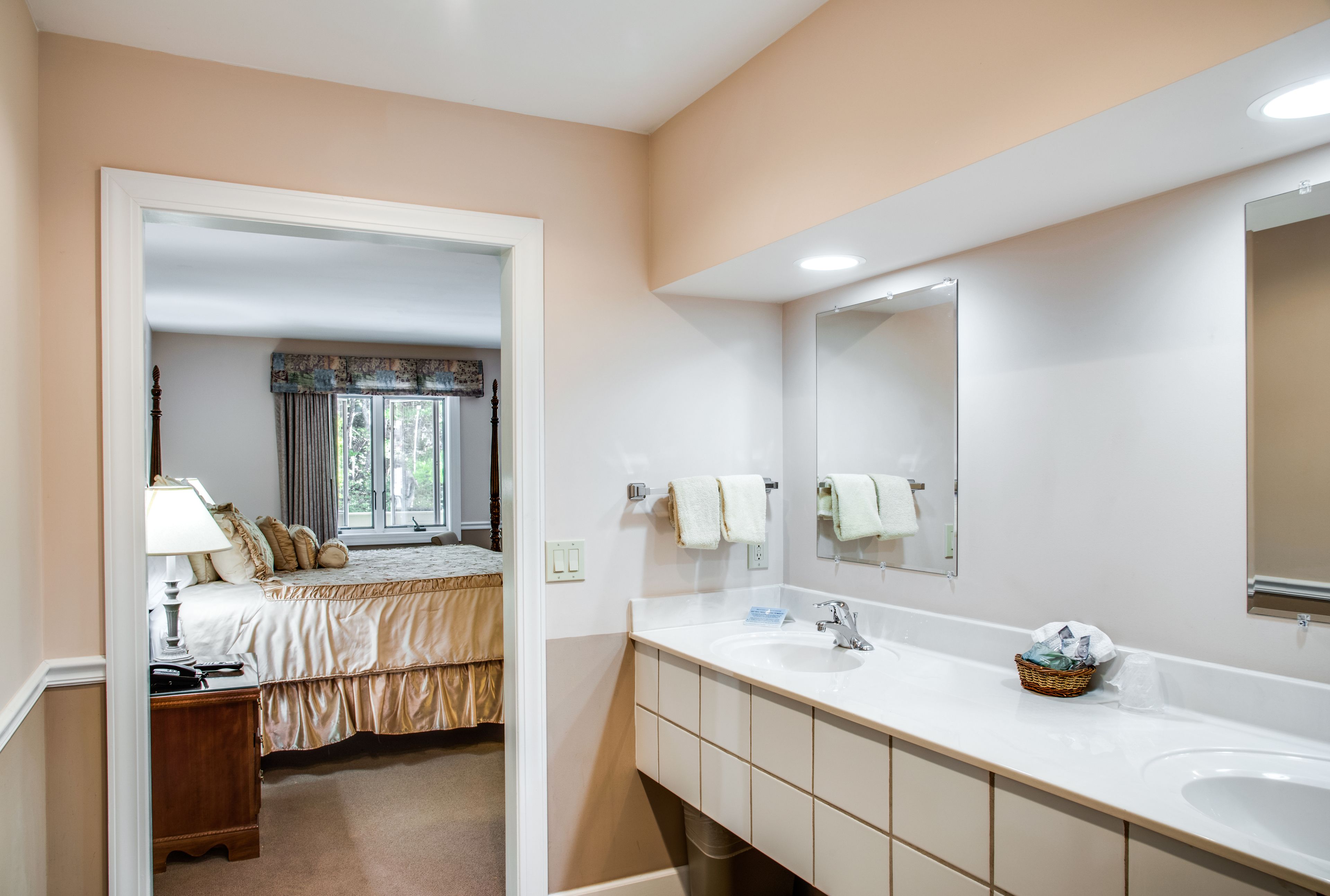 Ein Hotelbad mit einem Doppelwaschbecken, einem großen Spiegel und einem Bett, das durch eine offene Tür zu sehen ist.