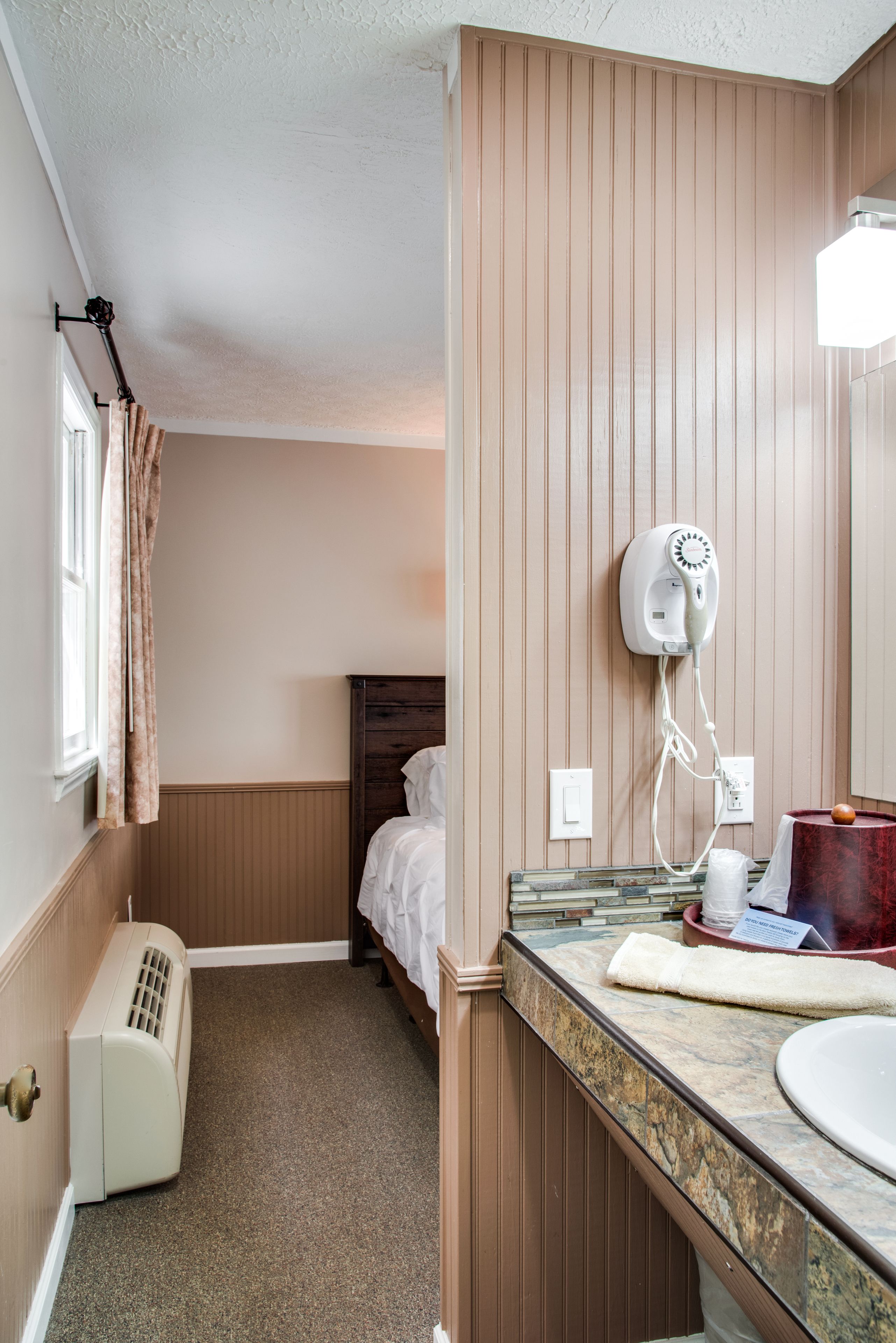 A hotel bathroom with a wooden vanity, a mirror, a hairdryer, and a window with a curtain.