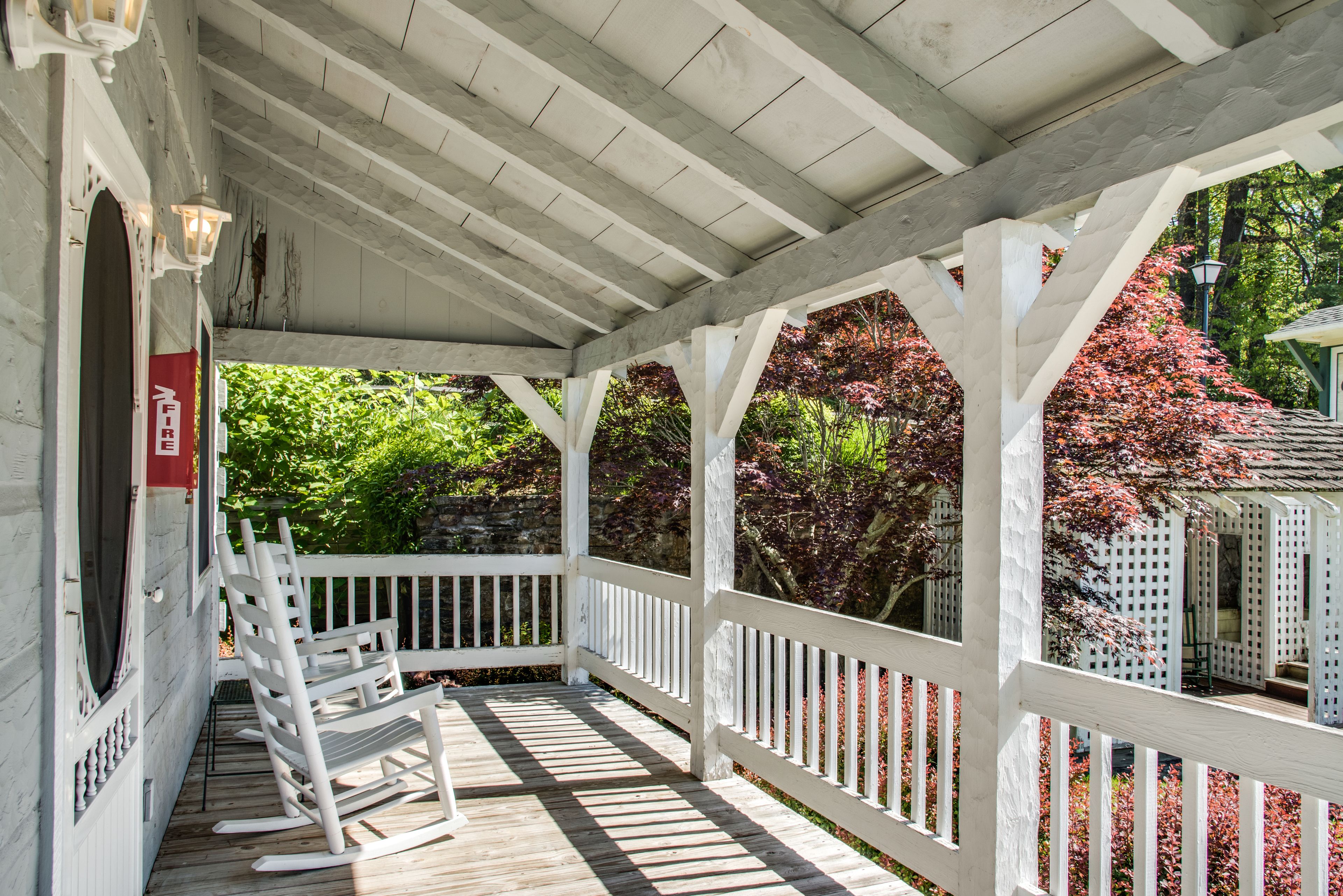 A covered porch with white railings and wooden flooring, surrounded by lush greenery and trees with red foliage.