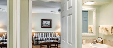 Un pasillo que conduce a una sala de estar con un sofá y un cuadro en la pared, y un baño con lavamanos y espejo.