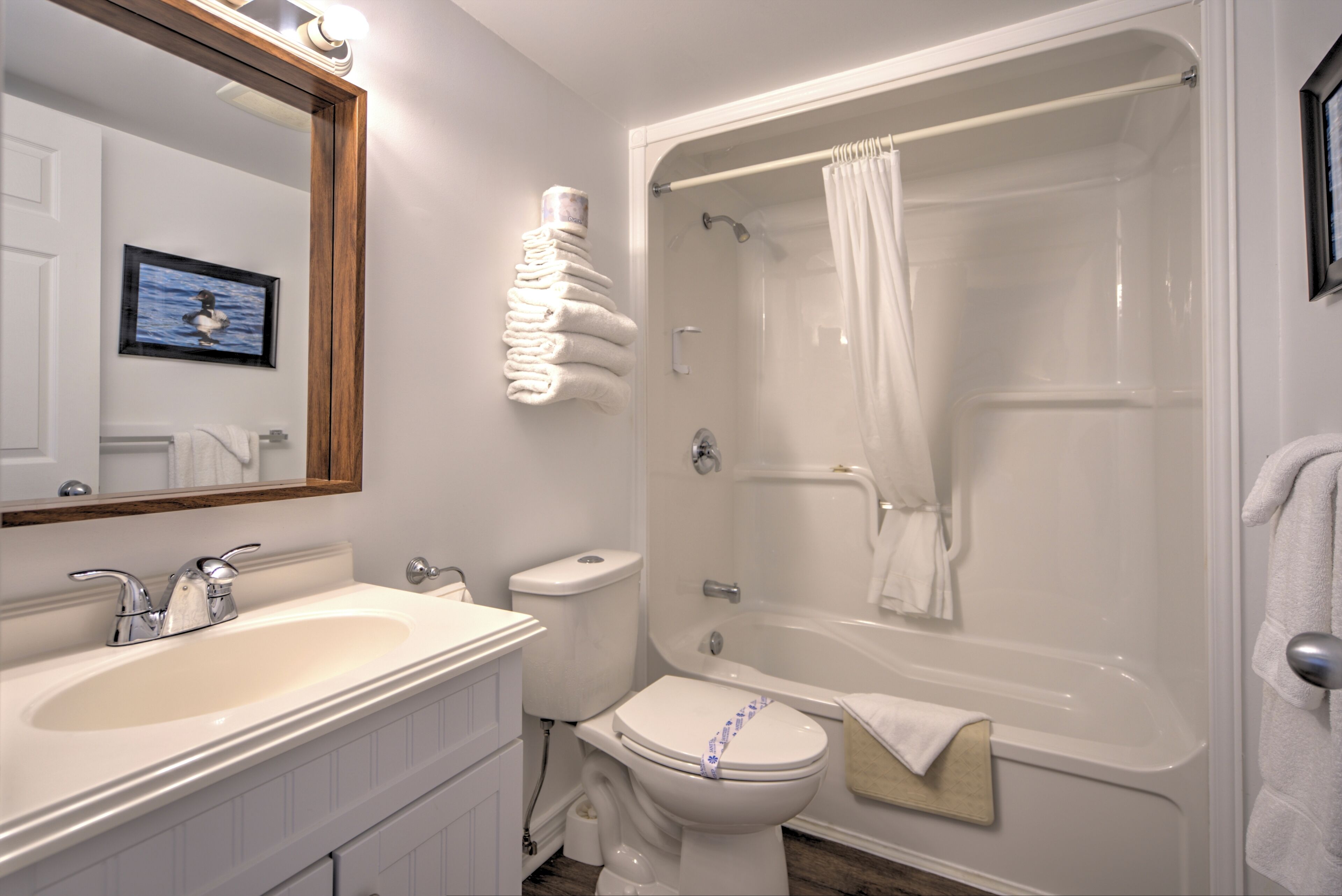 Ein Badezimmer mit einem weißen Waschbecken, einer Toilette und einer Badewanne. Handtücher sind an der Wand gestapelt und über dem Spiegel hängt ein gerahmtes Bild.