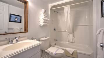 Ein Badezimmer mit einem weißen Waschbecken, einer Toilette und einer Badewanne. Handtücher sind an der Wand gestapelt und über dem Spiegel hängt ein gerahmtes Bild.