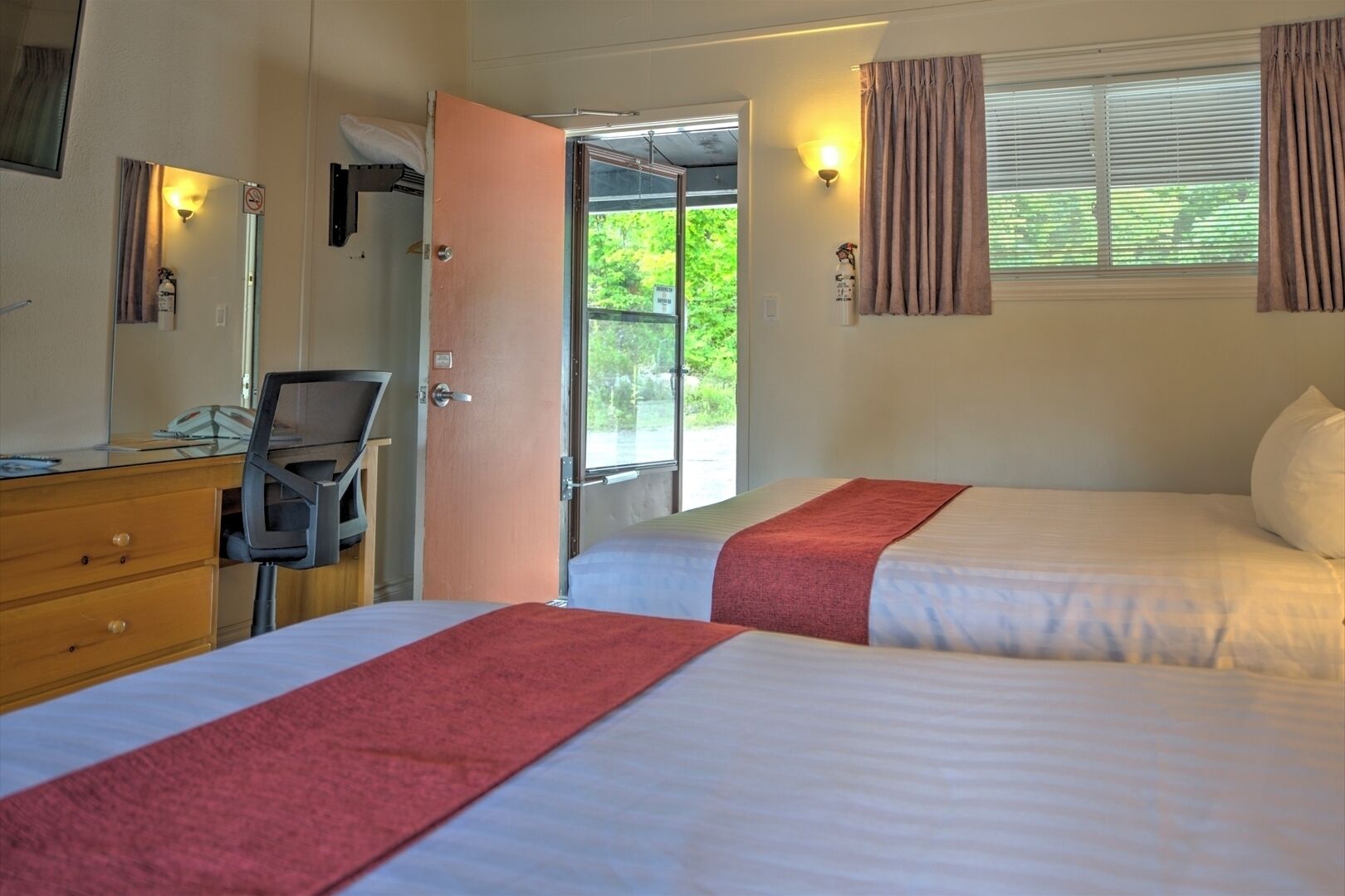 A hotel room with two beds, a desk with a chair, a mirror, and a window with curtains.