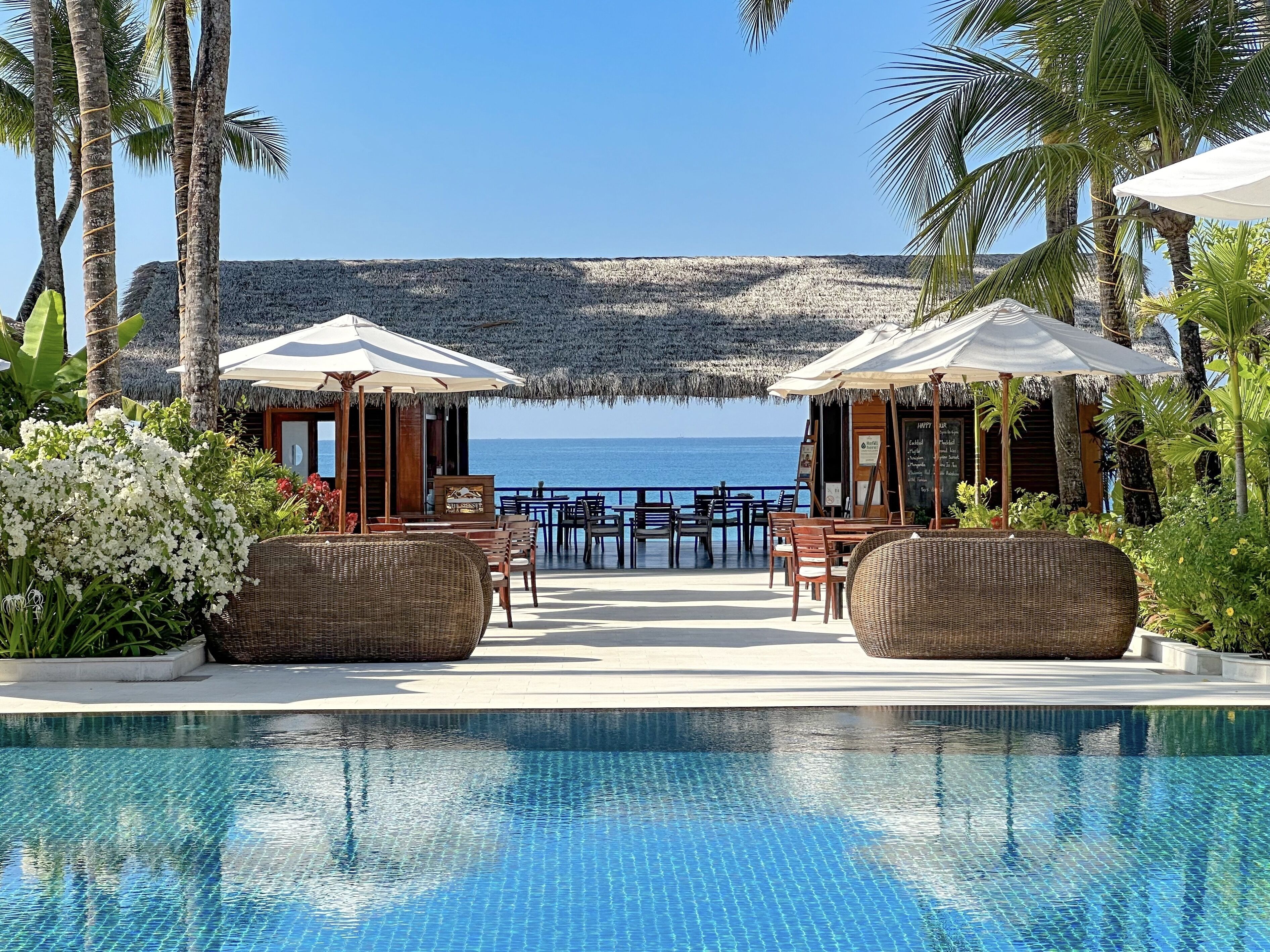 A poolside area with lounge chairs and umbrellas, overlooking a beach with a rocky outcrop in the distance.