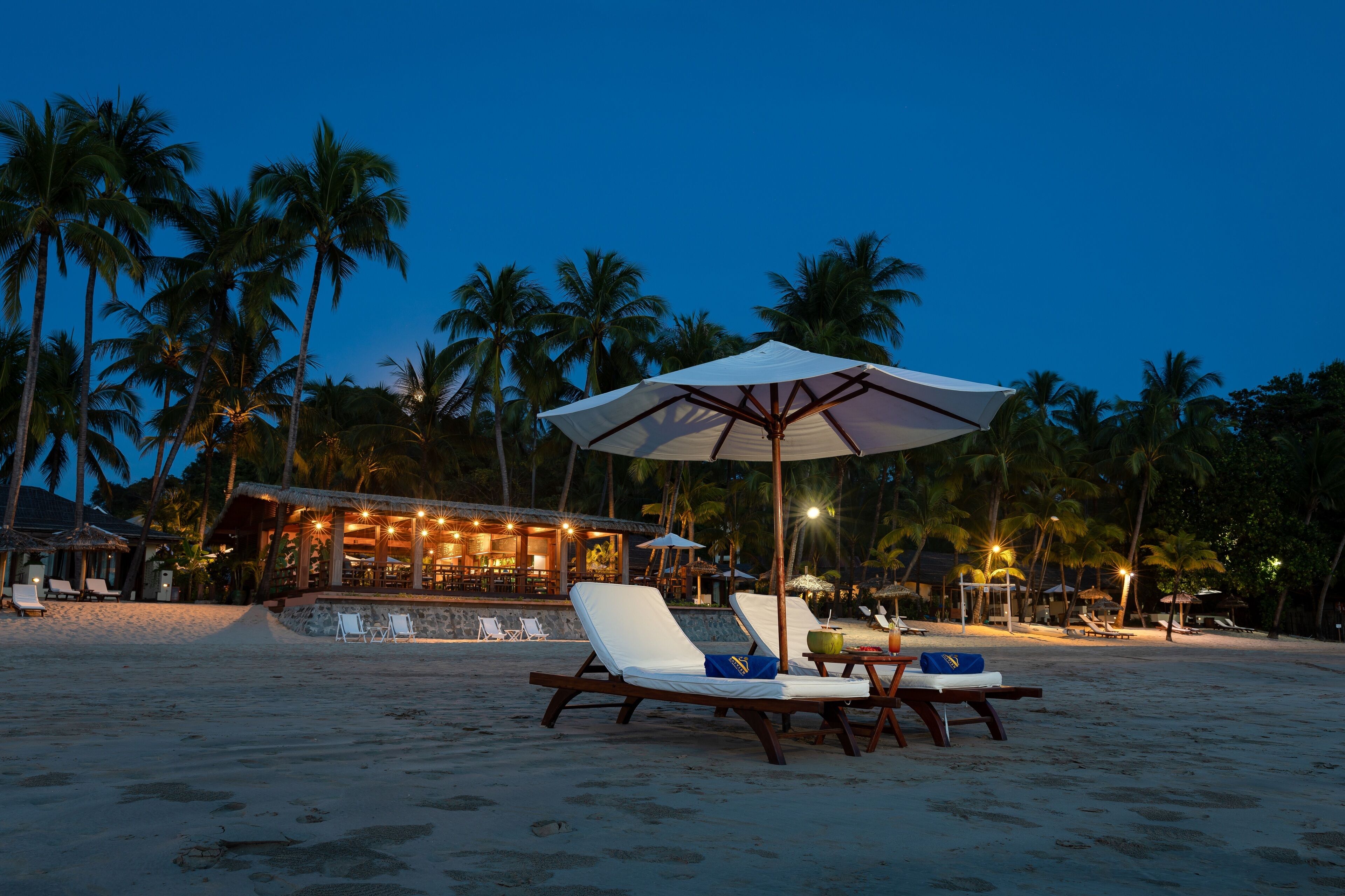 A beachside resort with lounge chairs under umbrellas, palm trees, and a bar area with ambient lighting.