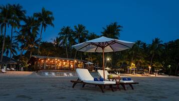 A beachside resort with lounge chairs under umbrellas, palm trees, and a bar area with ambient lighting.