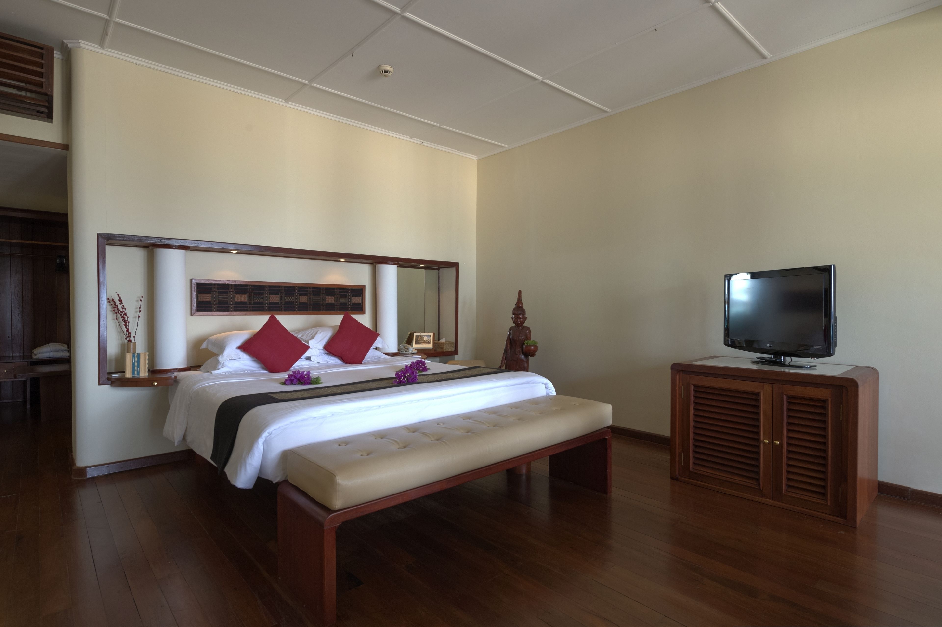 Habitación de hotel con una cama, un banco, un televisor sobre un mueble bajo y un espejo encima de la cama.