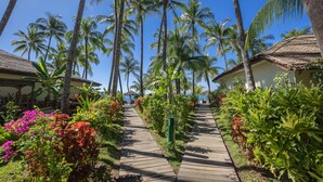 A wooden pathway flanked by lush greenery and palm trees, leading to a building with a thatched roof. - Bayview The Beach Resort (Ngapali)