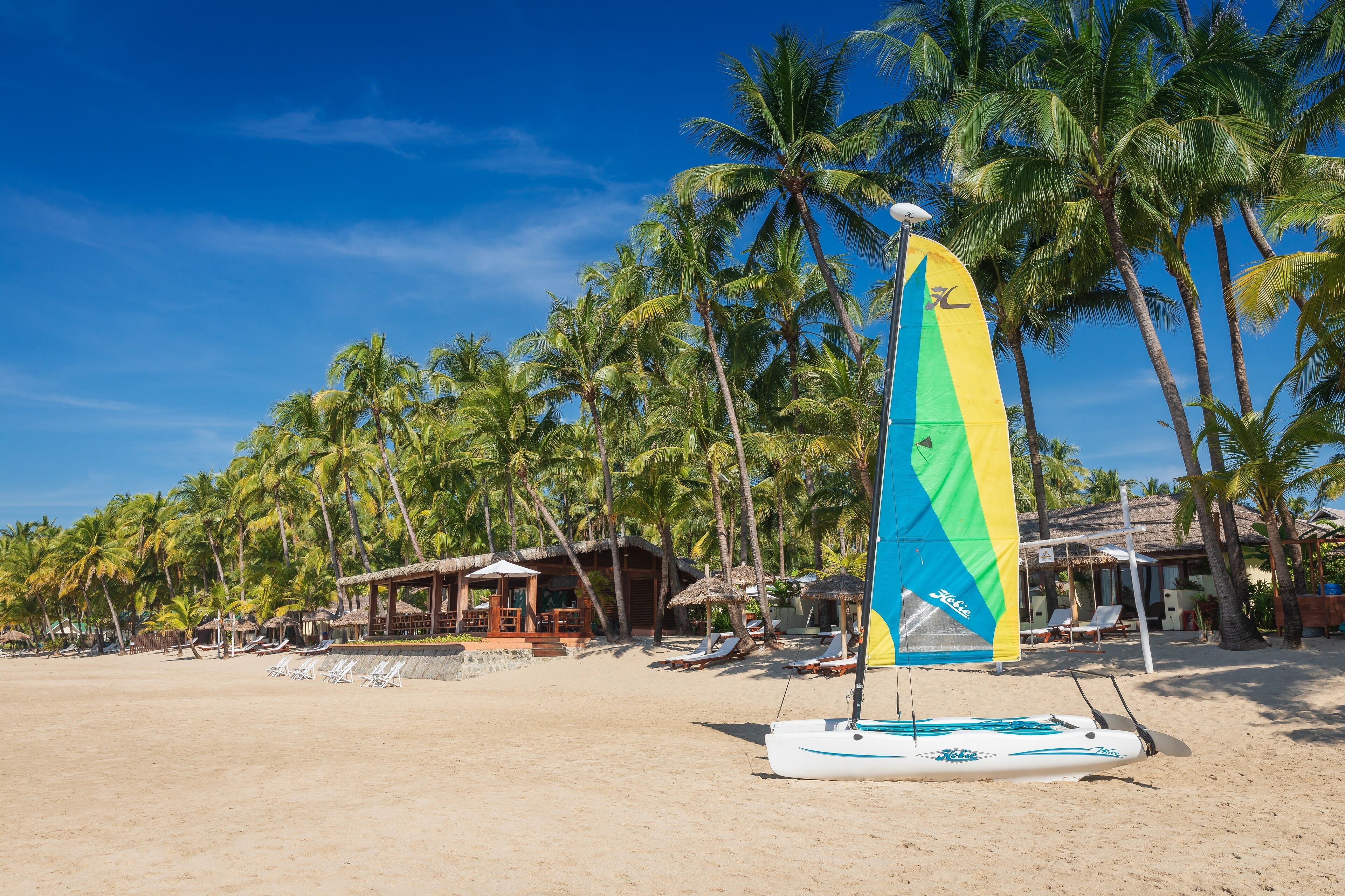Una playa con un velero, palmeras y cabañas.