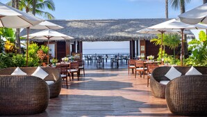 A wooden deck with wicker furniture and umbrellas, overlooking a beach with palm trees. - Bayview The Beach Resort (Ngapali)