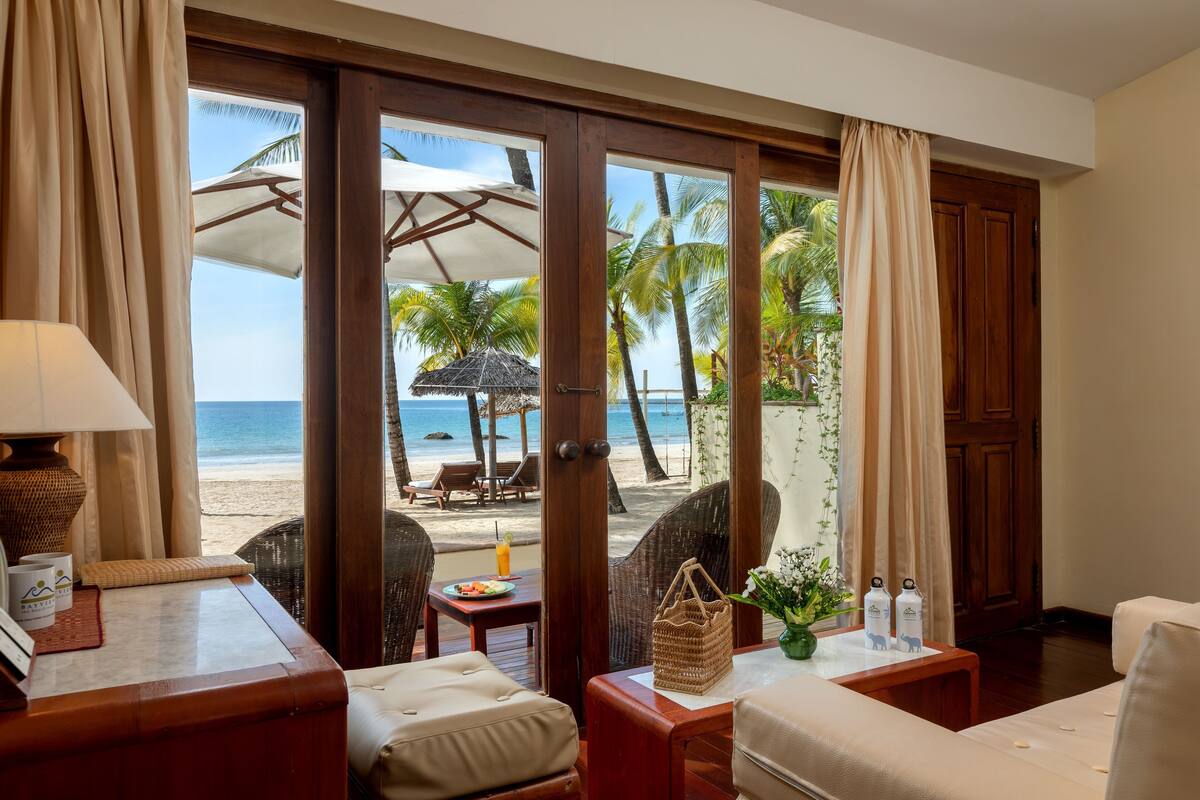 A room with a view of the beach, featuring a sofa, a table with a drink, and a wicker chair.