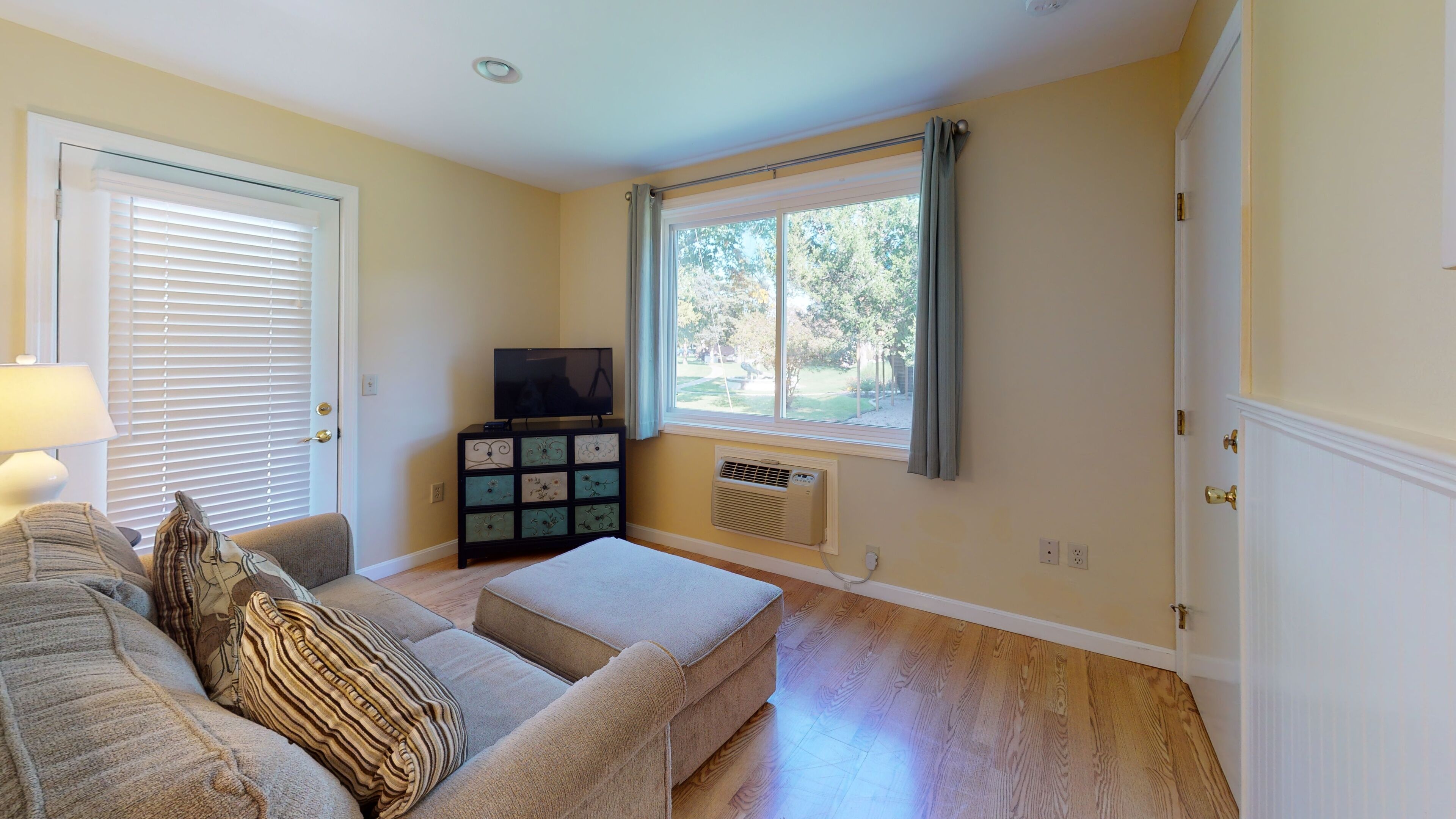 Un salón con sofá, reposapiés, mueble para televisor y una ventana con cortinas.
