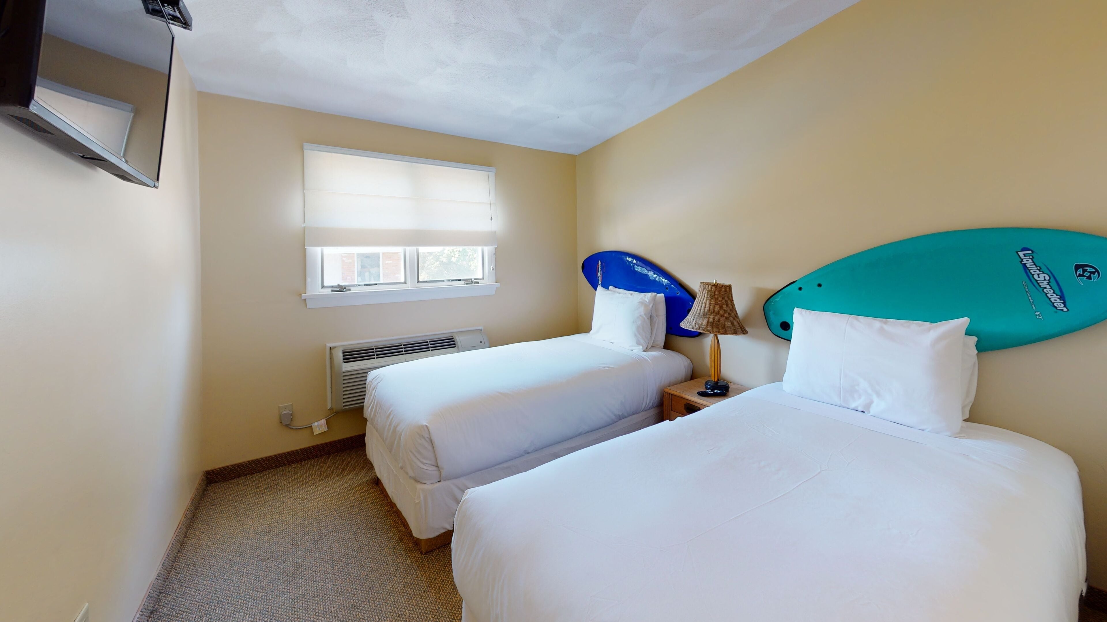 A hotel room with two beds, a window with blinds, a ceiling fan, and a surfboard on the wall.