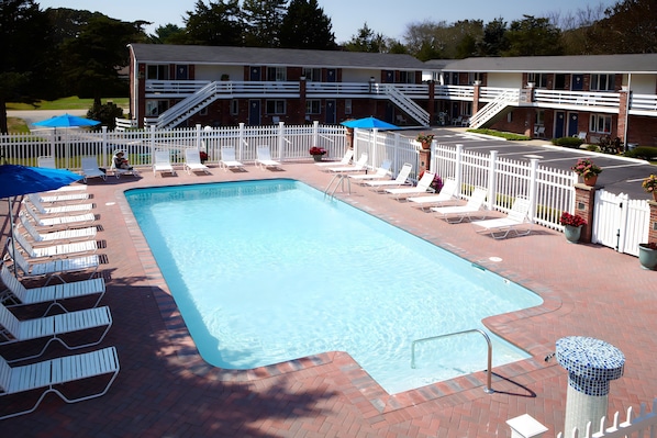 A rectangular pool with a white fence and lounge chairs around it, a covered area with umbrellas, and a building with a brick facade in the background.