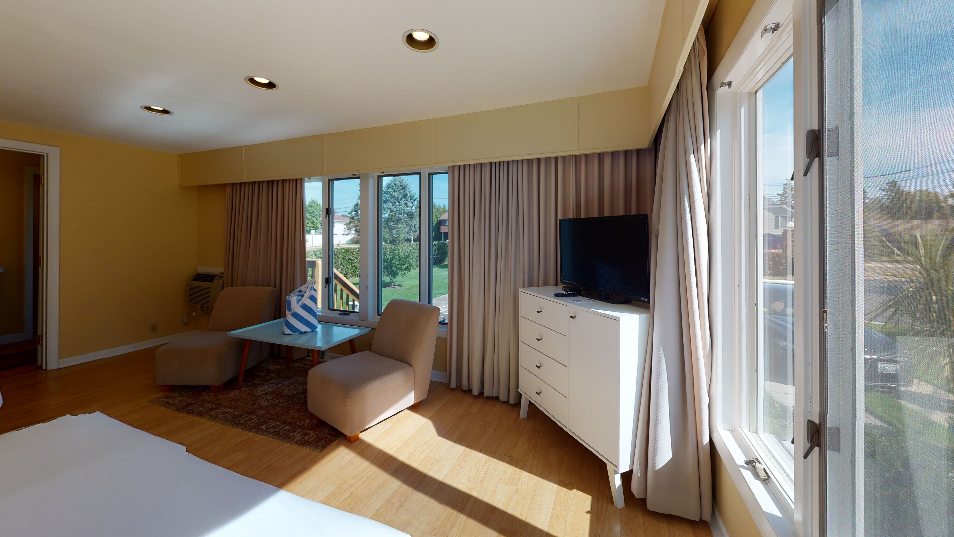 A modern hotel room with a large bed, a seating area with a table, a TV on a cabinet, and a view of the outdoors through large windows.
