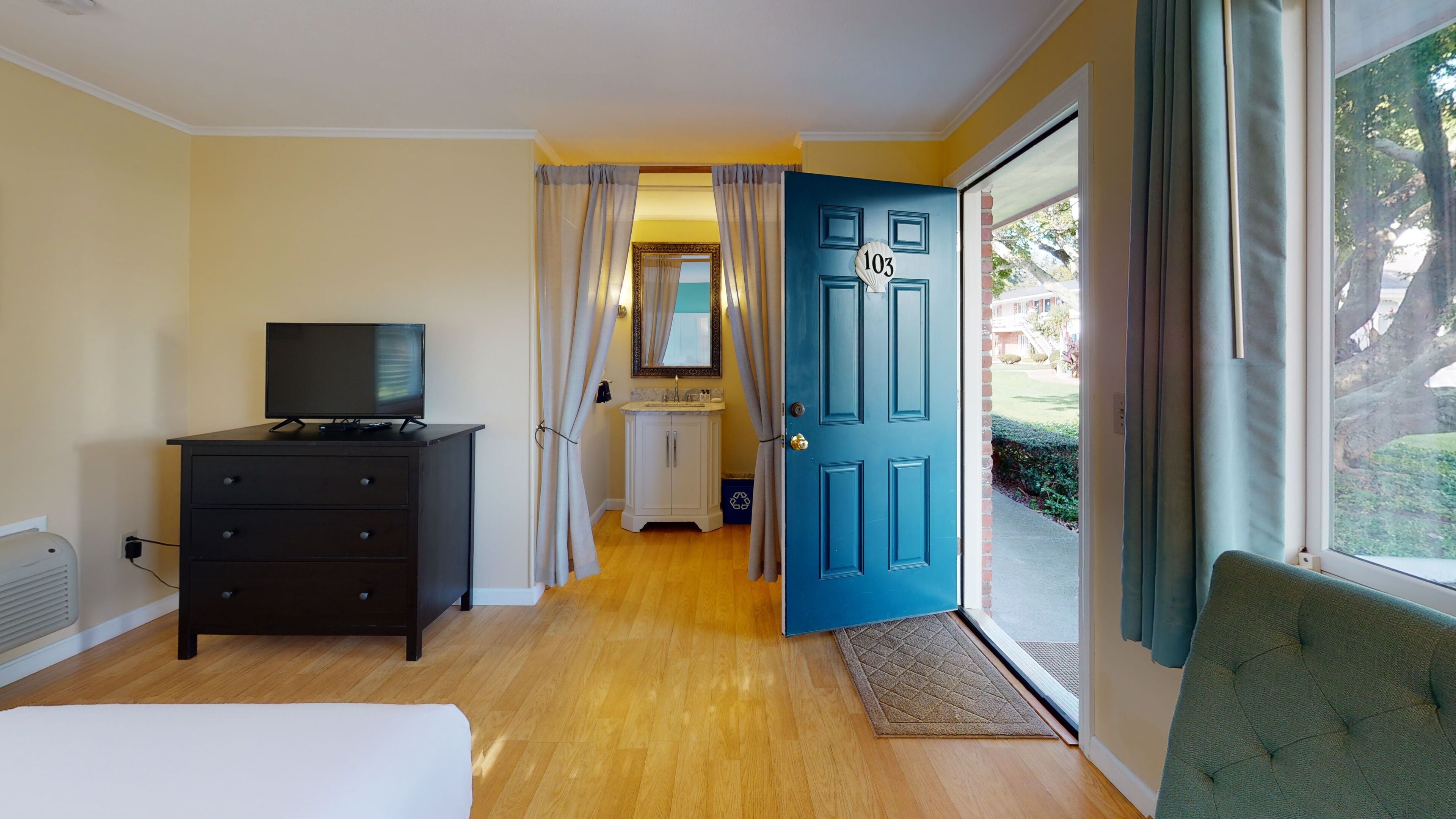 Habitación con televisión sobre una cómoda, cama, una puerta azul y vistas al exterior.