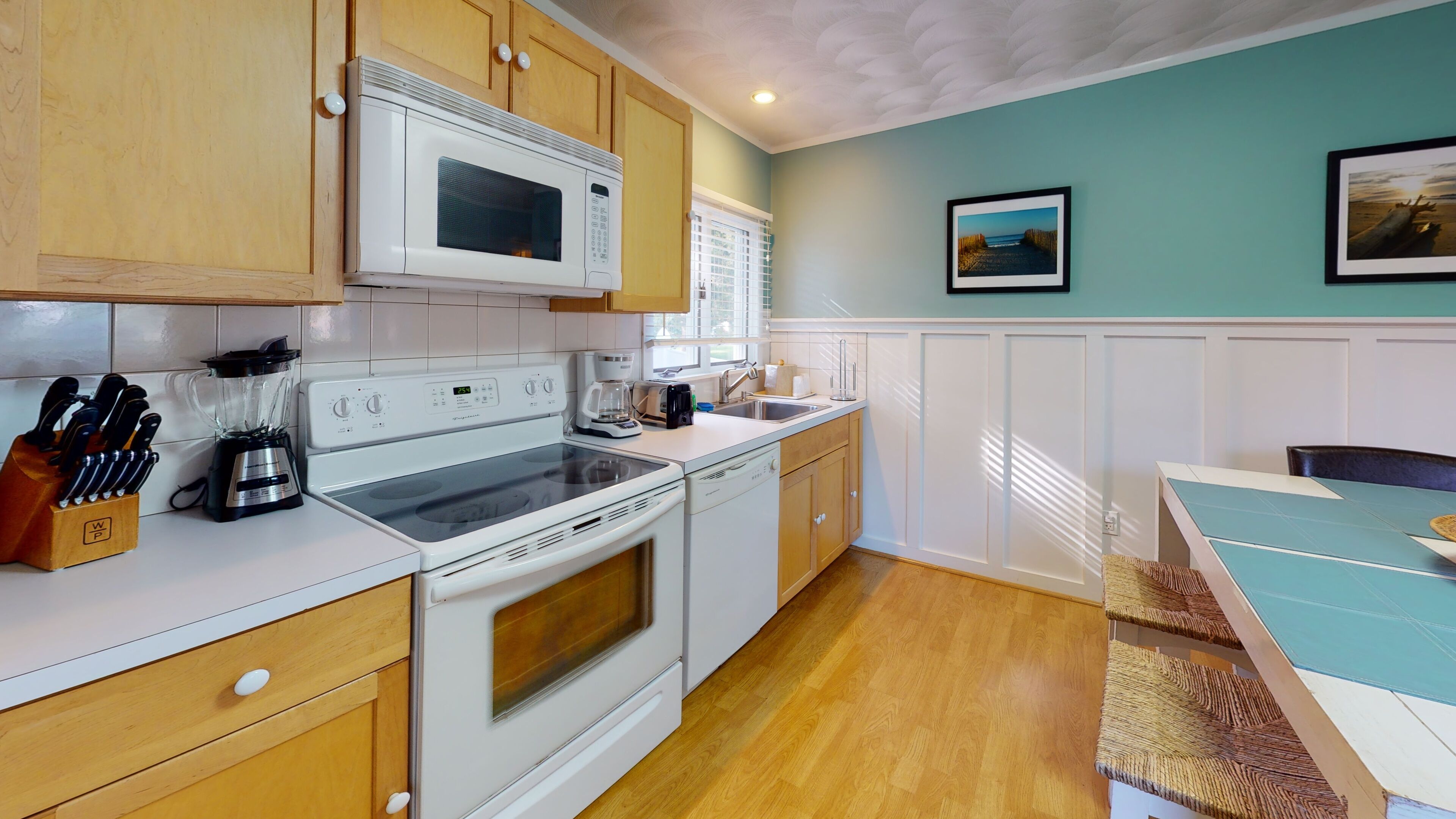 A kitchen with wooden cabinets, a microwave, stove, and a dishwasher. There is a window with blinds, a sink, and a countertop with a knife block and a blender.