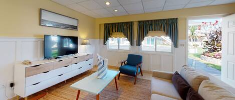 A living room with a flat-screen TV, a sofa, a chair, a small table, and a window with curtains.