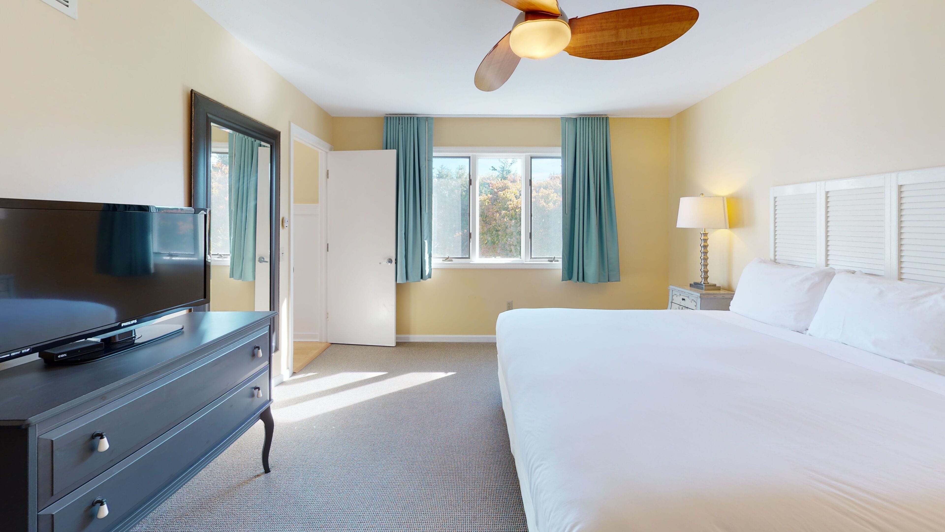 A hotel room with a bed, a television on a dresser, a ceiling fan, and a window with curtains.