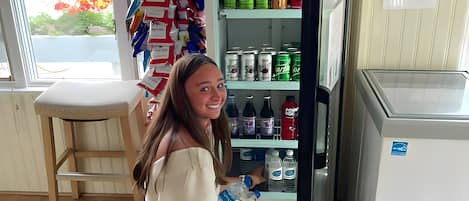 A person kneeling by a refrigerator stocked with beverages, a chair, and a window with a view of greenery.