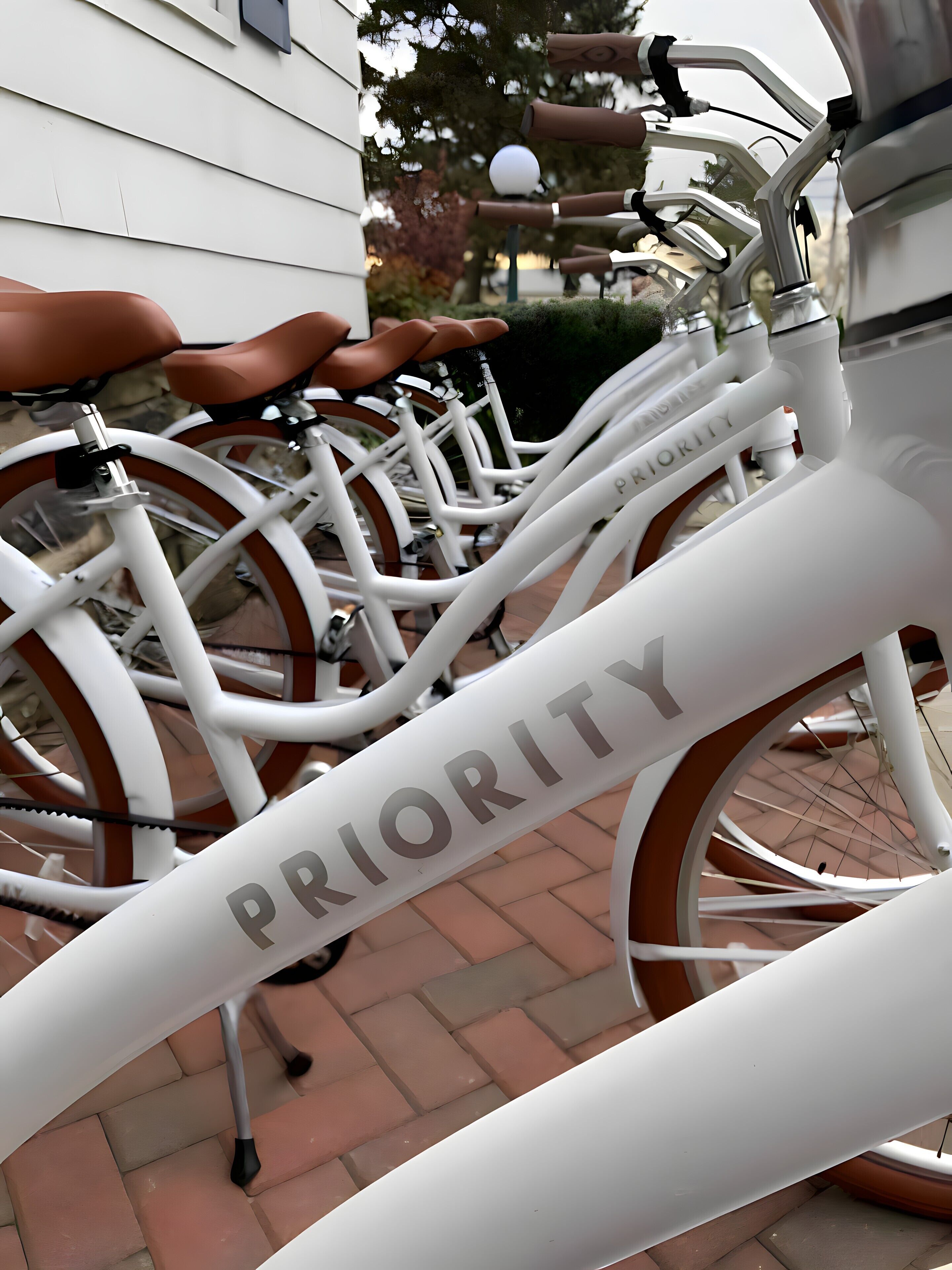 A white bicycle with the word "PRIORITY" on the frame, parked on a brick pavement.