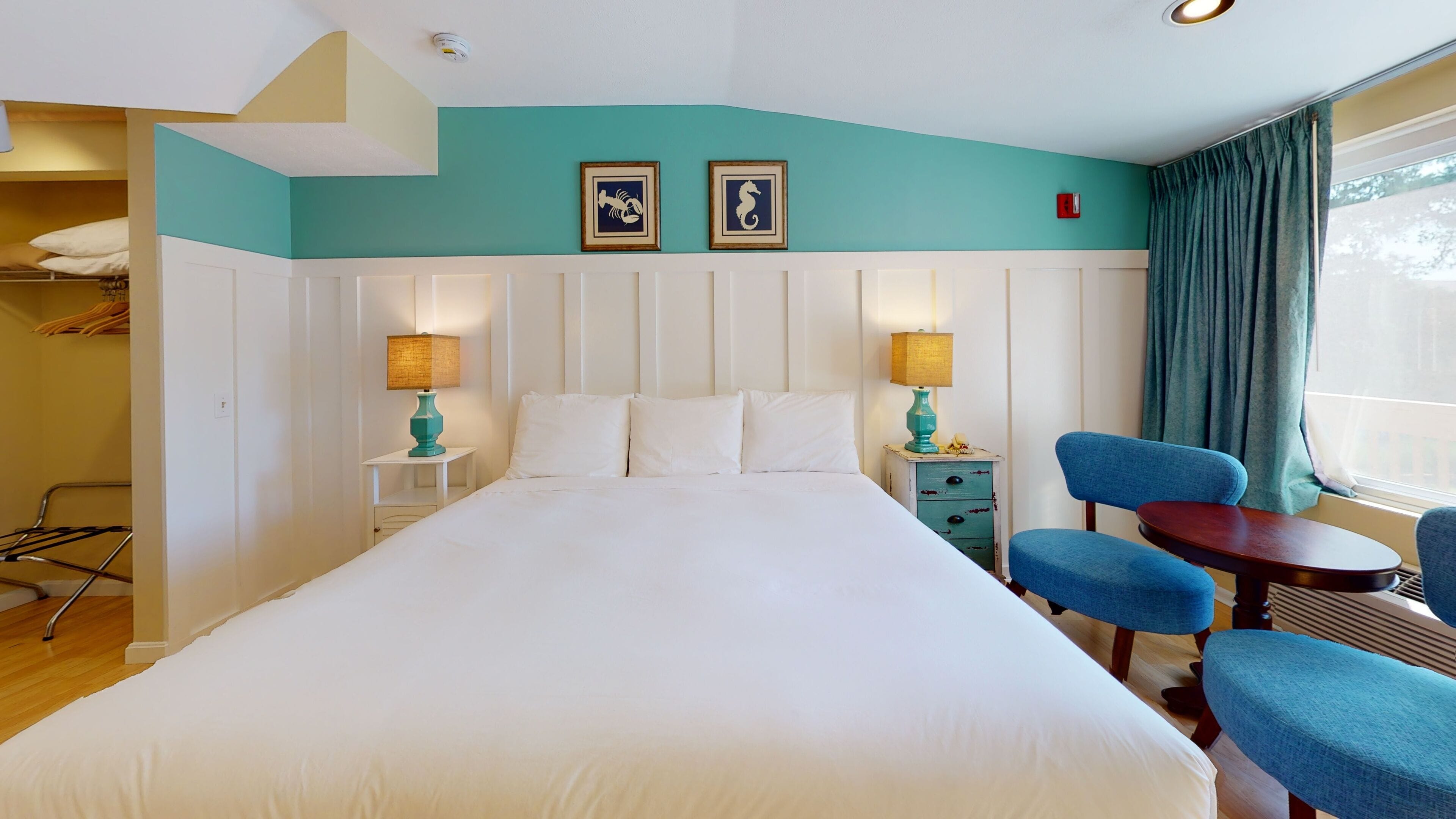 Une chambre d’hôtel avec un grand lit, deux tables de chevet avec des lampes, une chaise, une petite table et une fenêtre avec des rideaux.