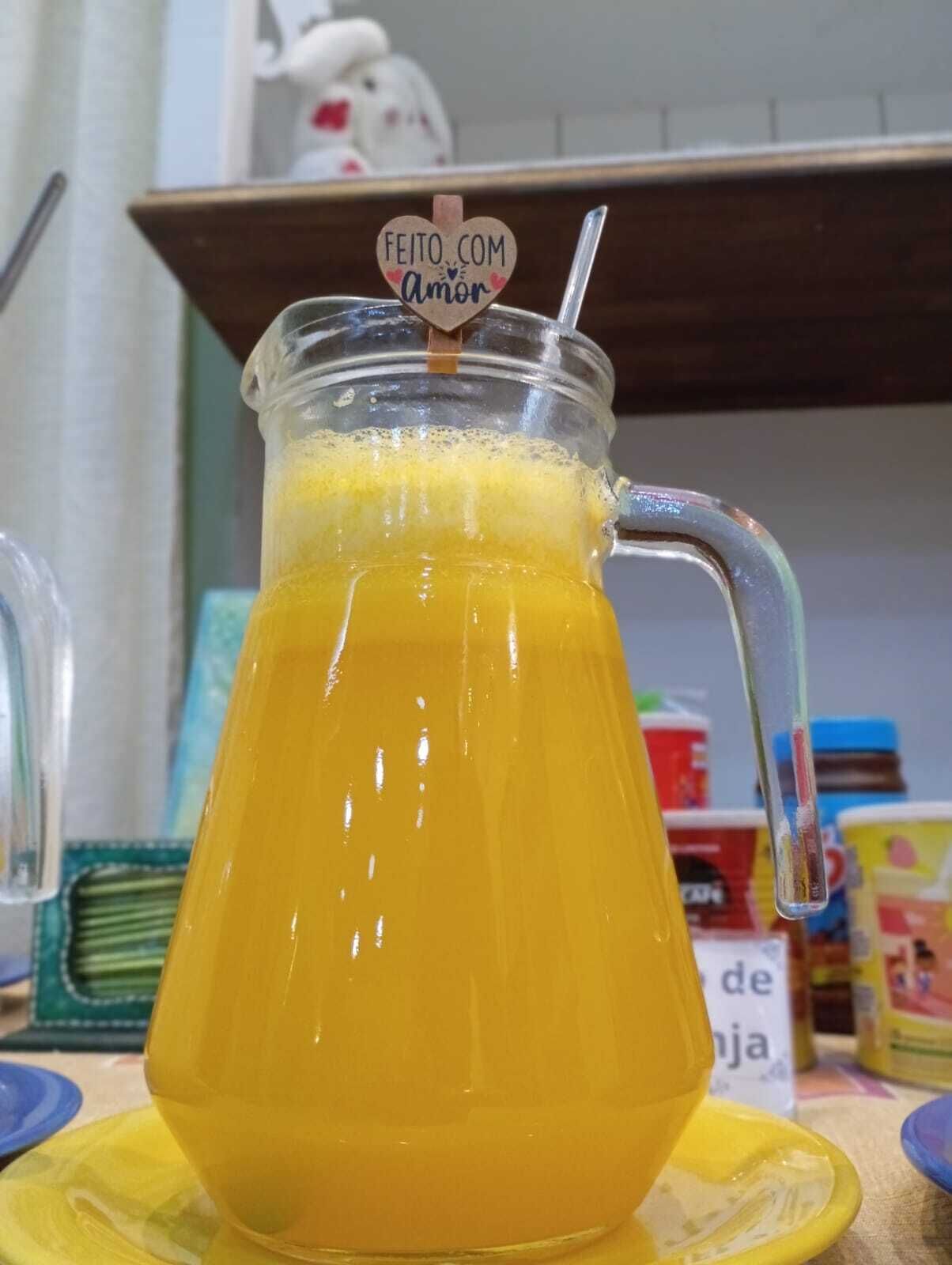 A glass pitcher filled with a yellow beverage, possibly lemonade, with a heart-shaped label that reads "FEITO COM Amor".