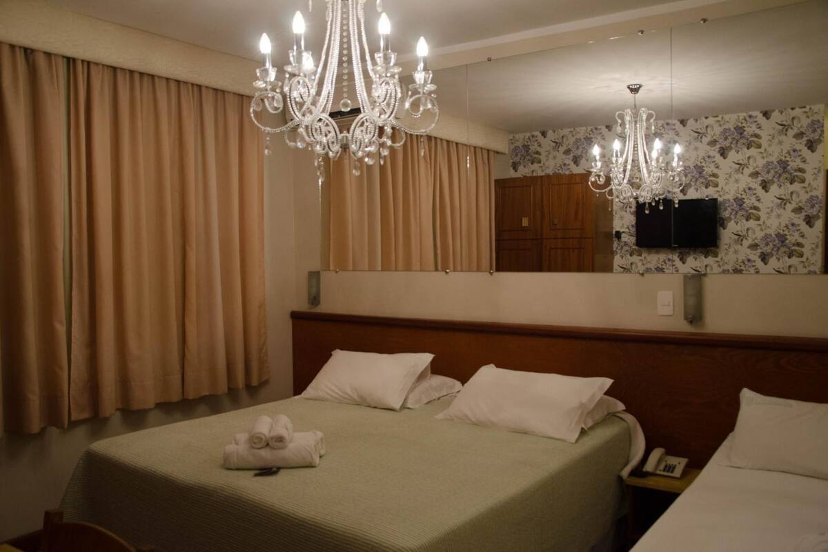 Una habitación de hotel con una cama grande, dos mesitas de noche, una araña, un televisor y un armario de madera.