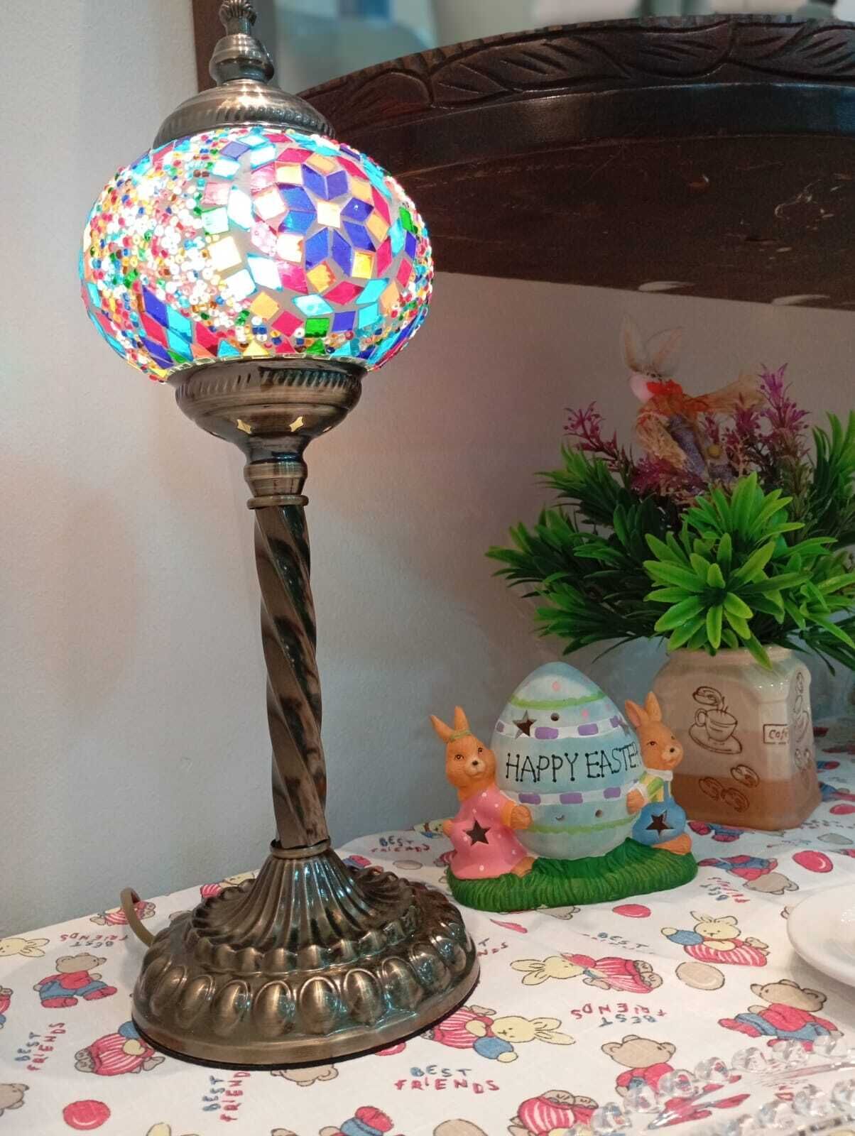 Una lámpara decorativa con un globo de mosaico, una figurita de conejo de cerámica y un huevo de Pascua de cerámica sobre una mesa con mantel estampado.