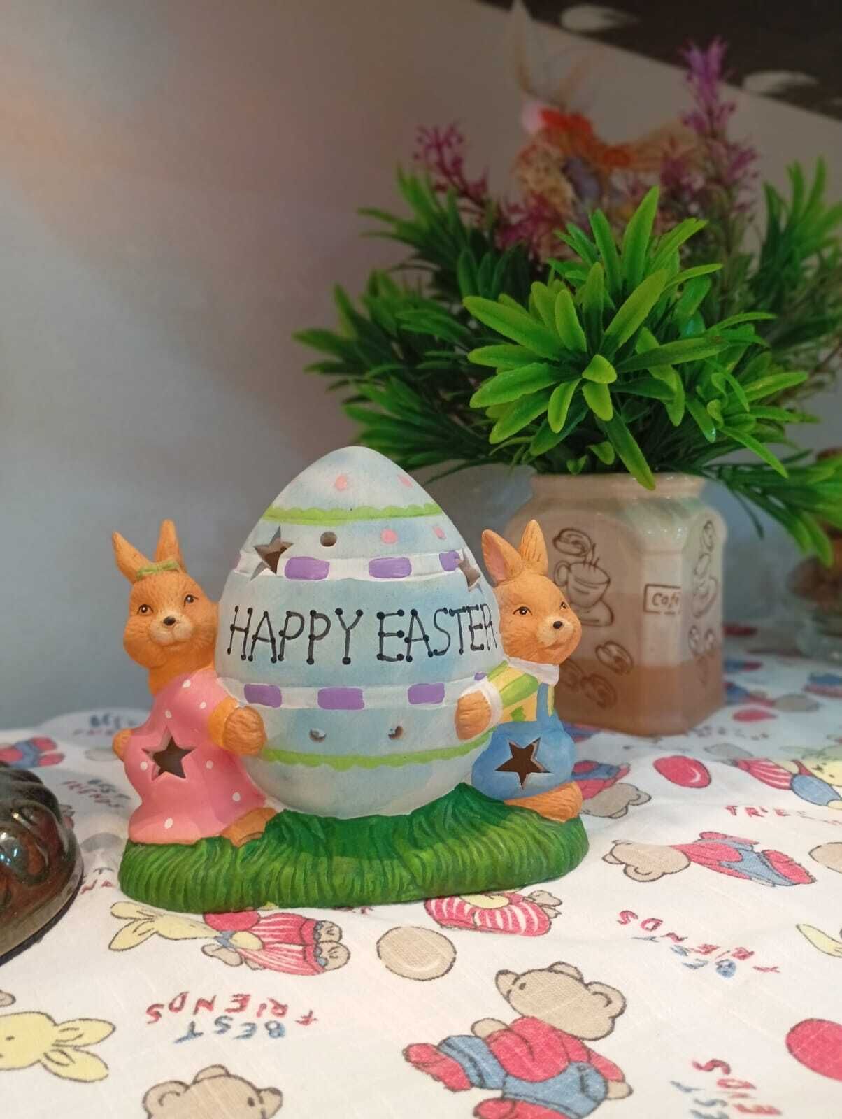 Ein dekoratives Osterszenario mit zwei Kaninchen und einem bemalten Ei auf einer Tischdecke mit Zeichentrickfiguren.